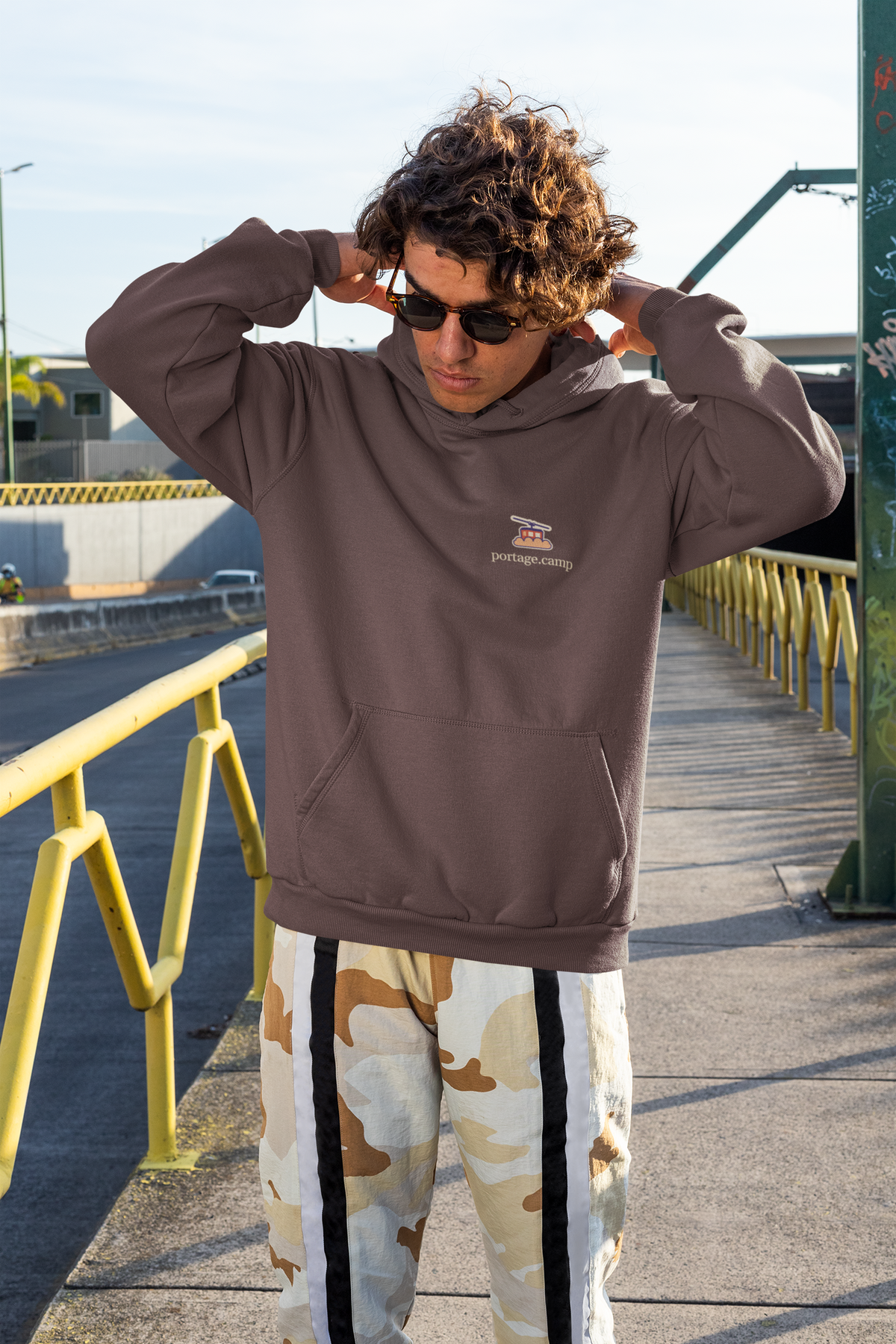 Cloud Ferry Heavyweight Hoodie in Brown, worn by a man on a city street, with the Cloud Ferry logo and portage.camp on the left chest. Digital product mockup.