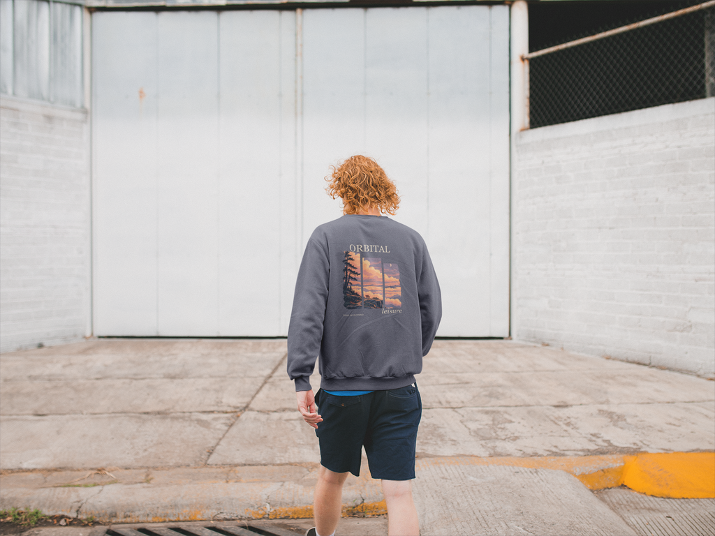 Edge of Evening Garment-Dyed Crewneck in Denim, worn by a man seen from behind walking away across an empty yard, showing the large print on the back. Digital product mockup.
