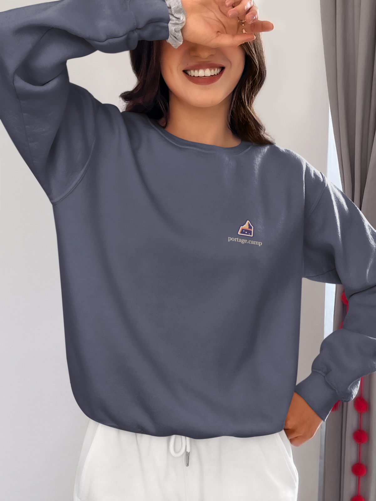 A Different Sky Garment-Dyed Crewneck in Denim, worn by a woman at home, with the A Different Sky logo and portage.camp on the chest. Digital product mockup.