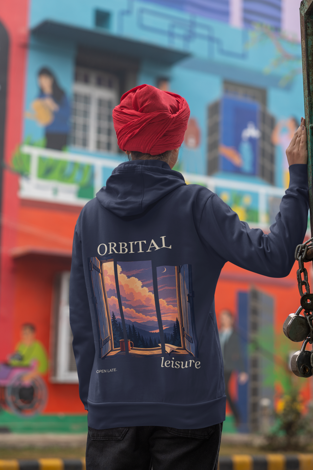 Open Late Zip Hoodie in Navy, worn by a woman seen from behind in a turban by a painted mural, showing the large print on the back. Digital product mockup.