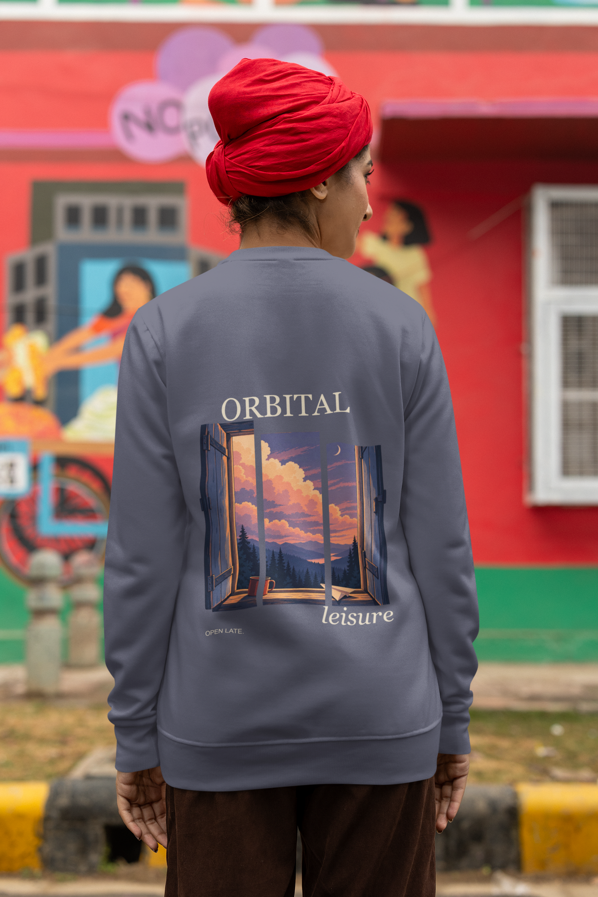 Open Late Garment-Dyed Crewneck in Denim, worn by a woman seen from behind in a turban by a painted mural, showing the large print on the back. Digital product mockup.