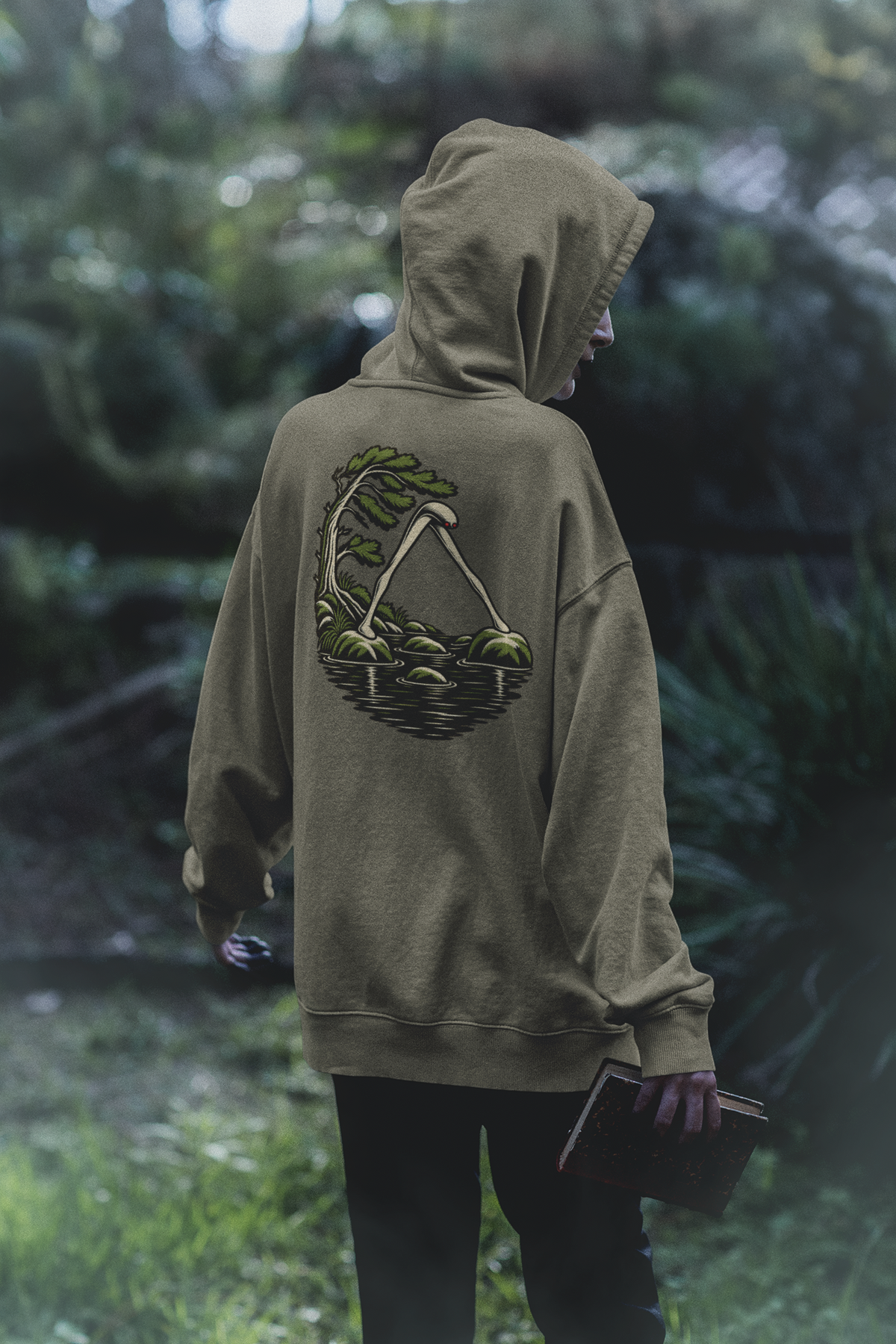 Small Hours Heavyweight Hoodie in Olive, worn by a woman seen from behind in a dark forest, showing the large print on the back. Digital product mockup.
