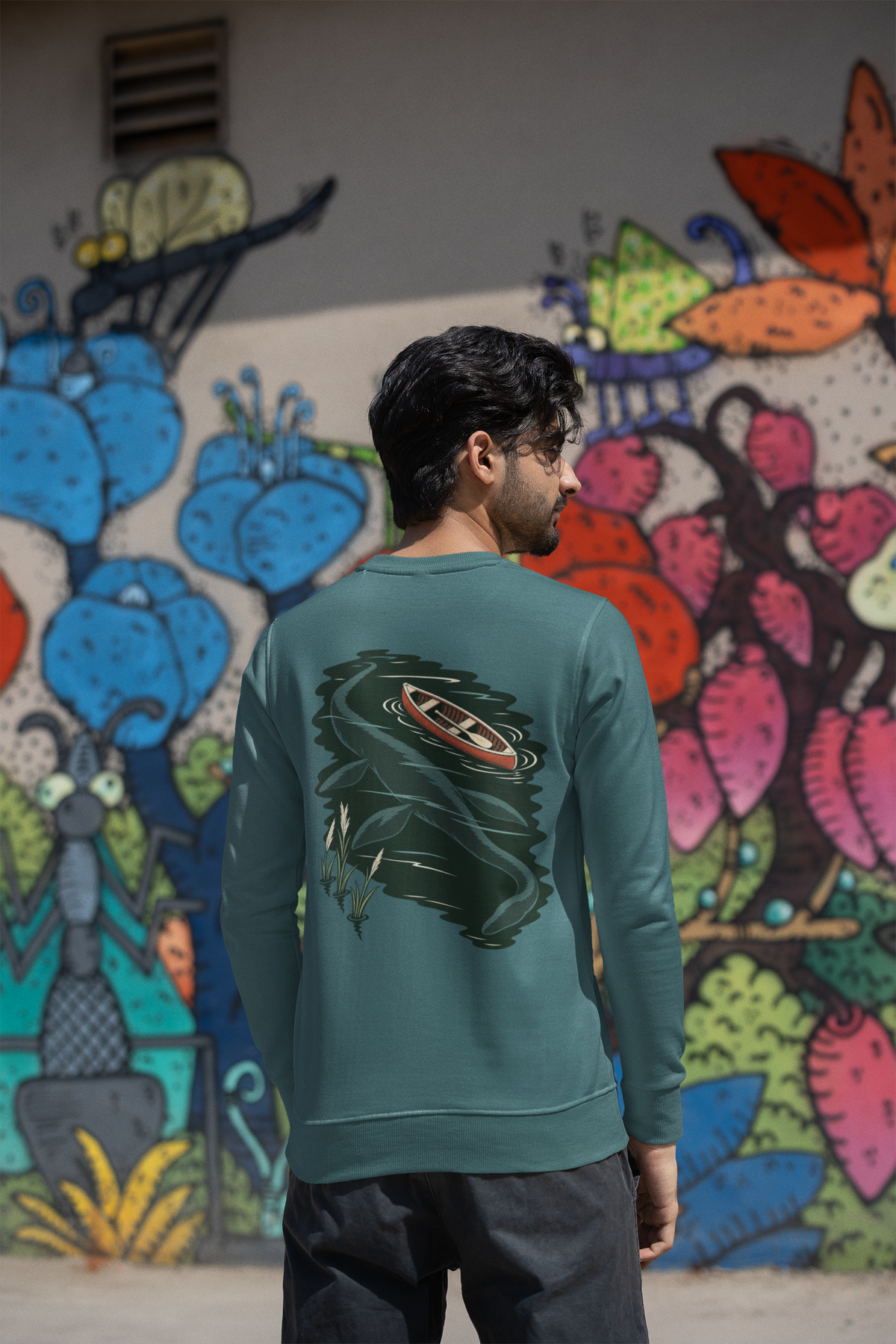 Seen Briefly Garment-Dyed Crewneck in Blue Spruce, worn by a man seen from behind by a painted wall in the city, showing the large print on the back. Digital product mockup.
