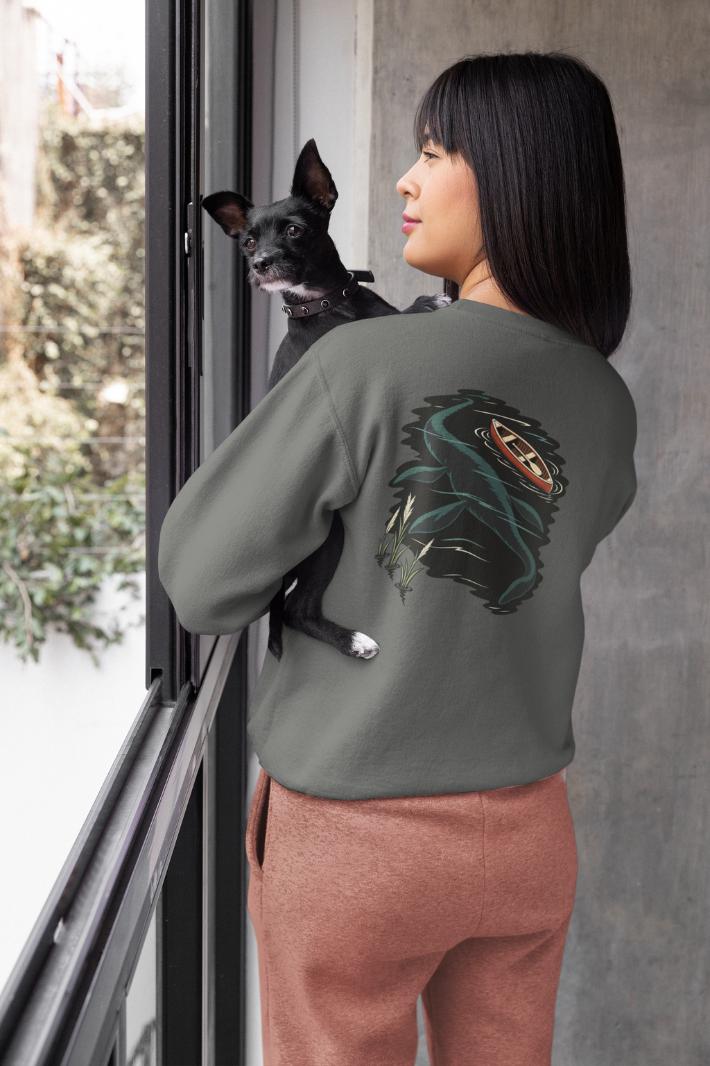 Seen Briefly Garment-Dyed Crewneck in Pepper, worn by a woman seen from behind at home, holding her dog, showing the large print on the back. Digital product mockup.