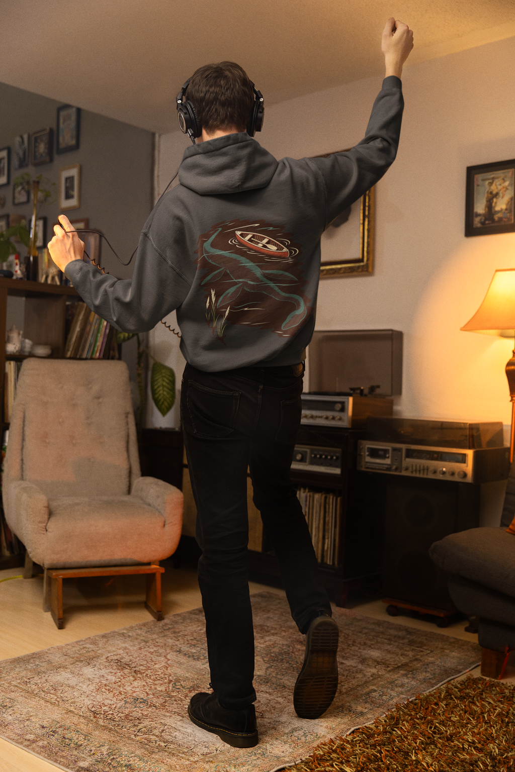 Small Hours Heavyweight Hoodie in Black, worn by a man seen from behind in a living room at night, showing the large print on the back. Digital product mockup.