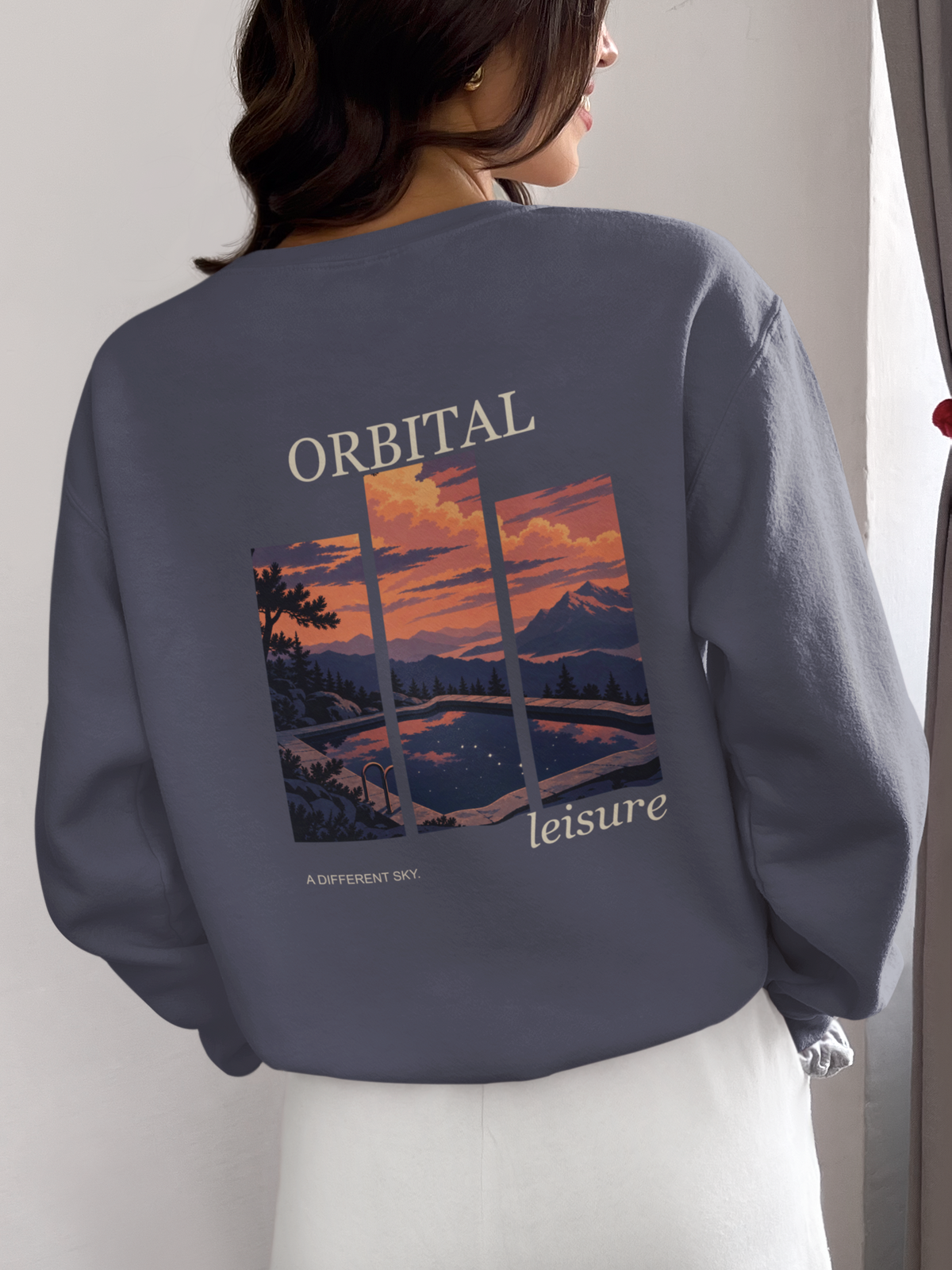 A Different Sky Garment-Dyed Crewneck in Denim, worn by a woman seen from behind at home, showing the large print on the back. Digital product mockup.