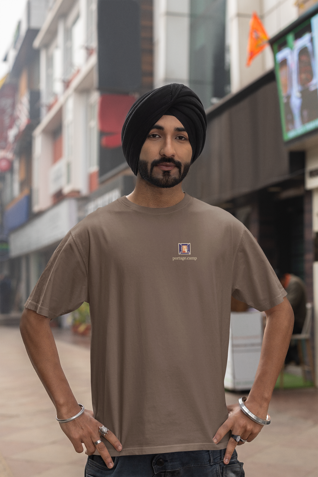 Open Late Garment-Dyed Tee in Espresso, worn by a man in a turban on a busy street, with the Open Late logo and portage.camp on the chest. Digital product mockup.