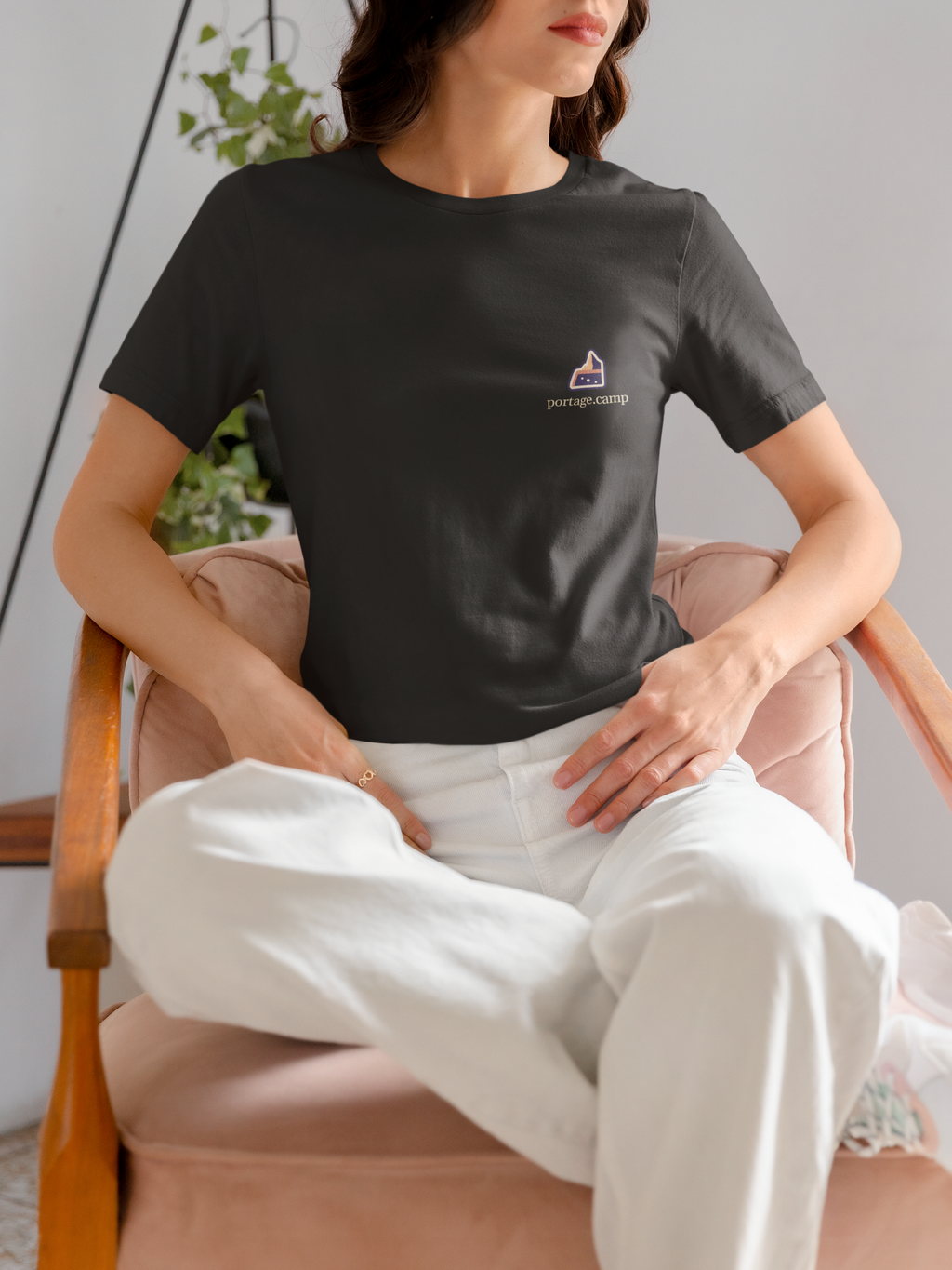 A Different Sky Garment-Dyed Tee in Black, worn by a woman seated on a chair at home, with the A Different Sky logo and portage.camp on the chest. Digital product mockup.