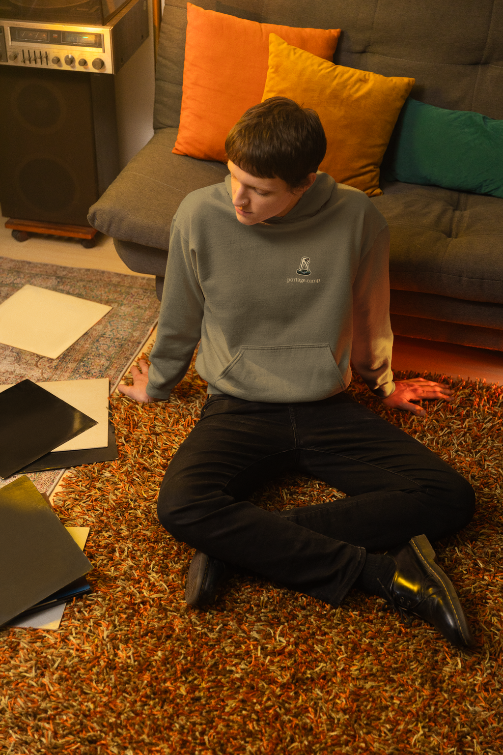 Small Hours Heavyweight Hoodie in Olive, worn by a man seated on the floor among records, with the Small Hours logo and portage.camp on the left chest. Digital product mockup.