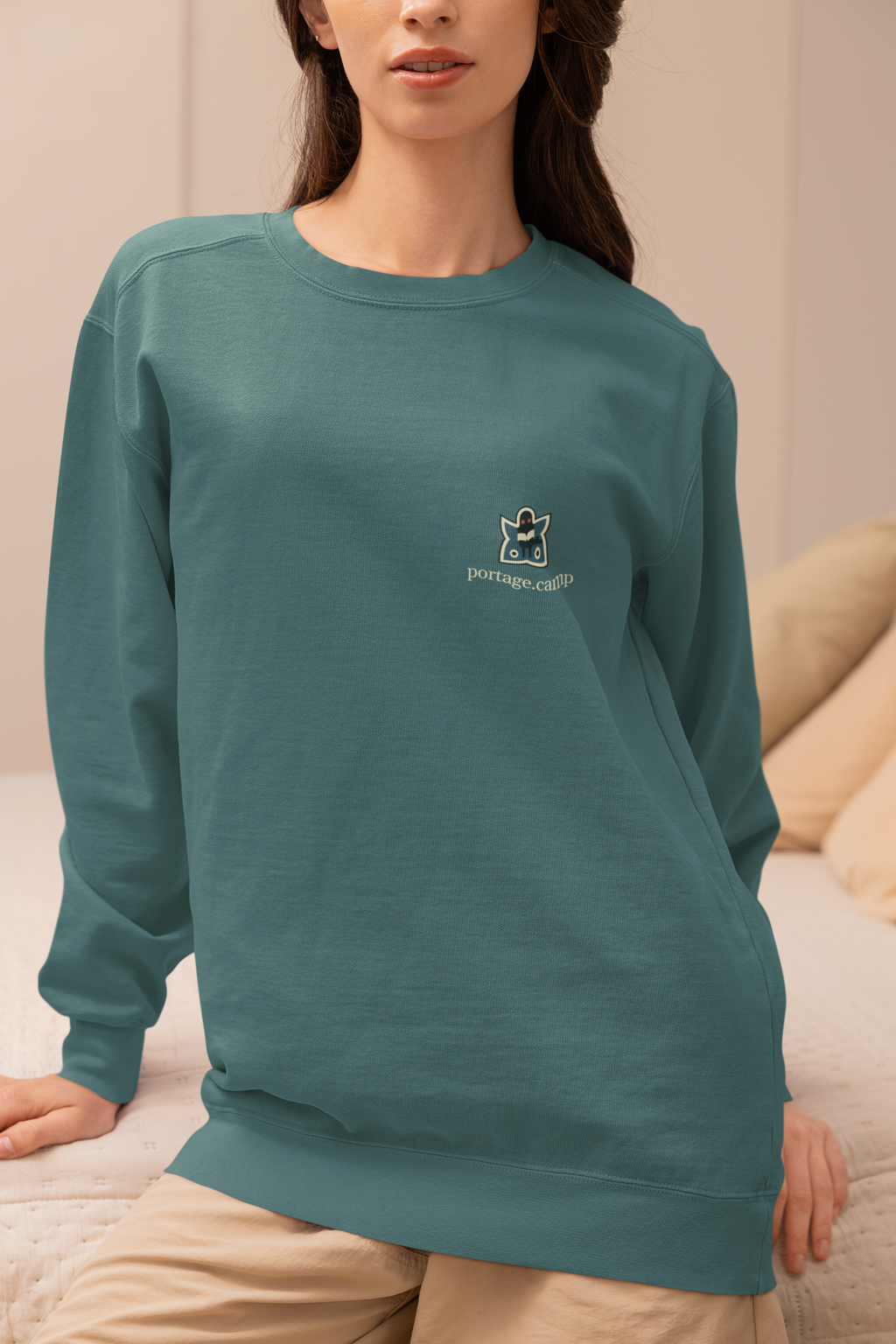 Night Library Garment-Dyed Crewneck in Blue Spruce, worn by a woman seated on a bed at home, with the Night Library logo and portage.camp on the chest. Digital product mockup.