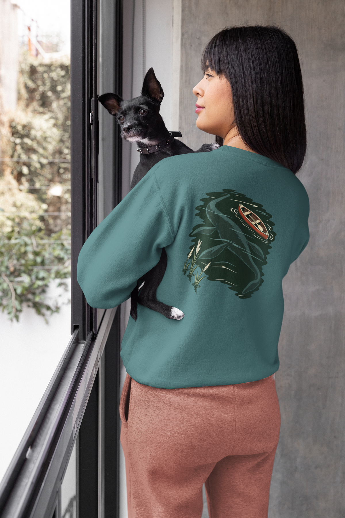 Seen Briefly Garment-Dyed Crewneck in Blue Spruce, worn by a woman seen from behind at home, holding her dog, showing the large print on the back. Digital product mockup.