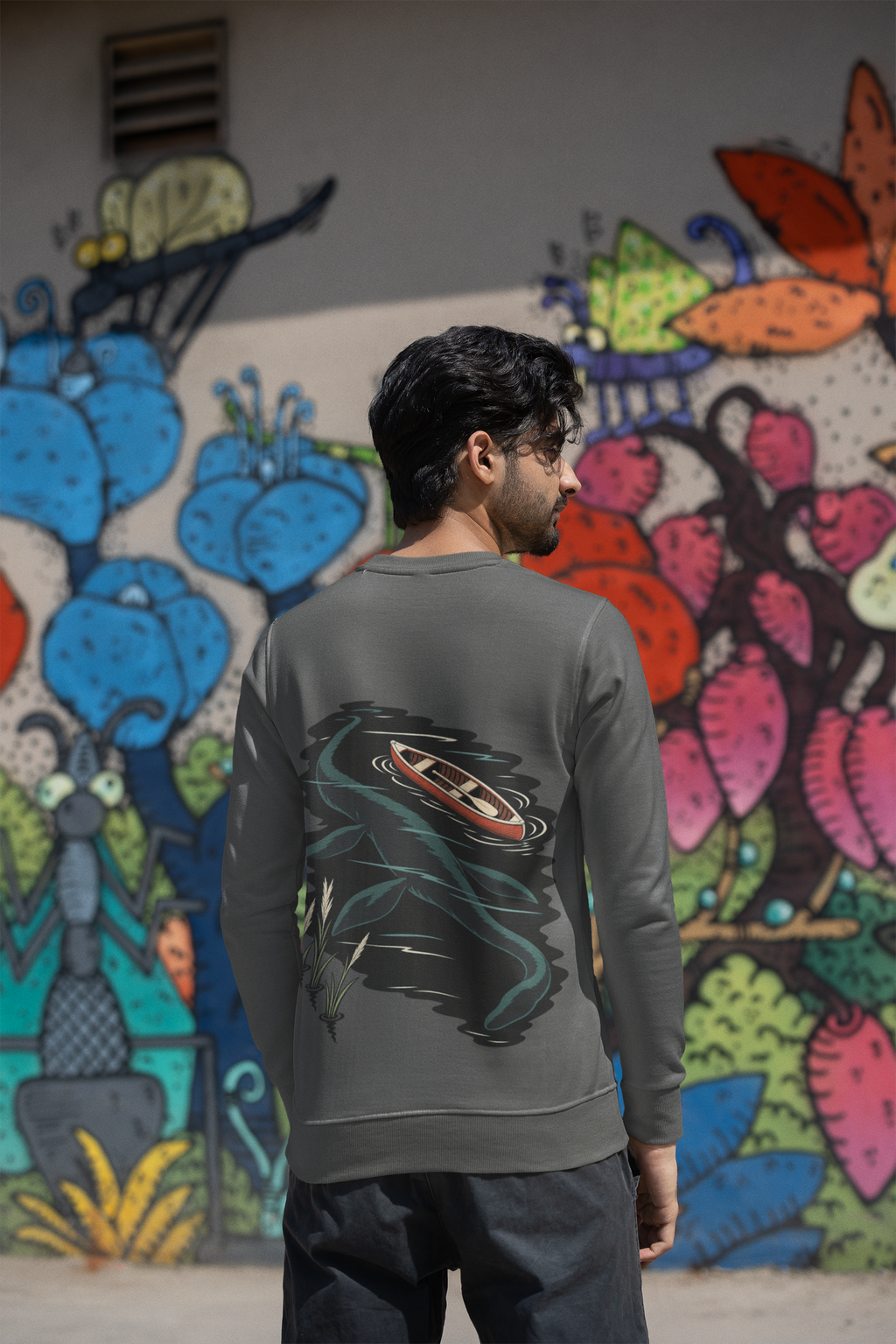 Seen Briefly Garment-Dyed Crewneck in Pepper, worn by a man seen from behind by a painted wall in the city, showing the large print on the back. Digital product mockup.