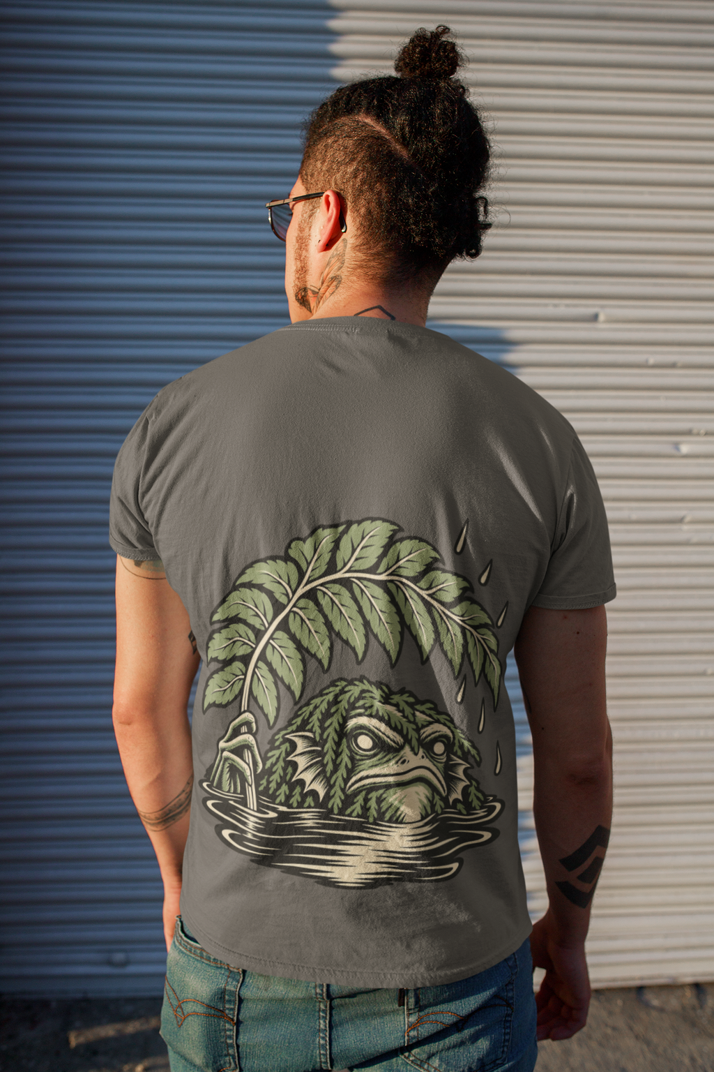 Rain Check Garment-Dyed Tee in Pepper, worn by a man seen from behind on a street beside a metal shutter, showing the large print on the back. Digital product mockup.
