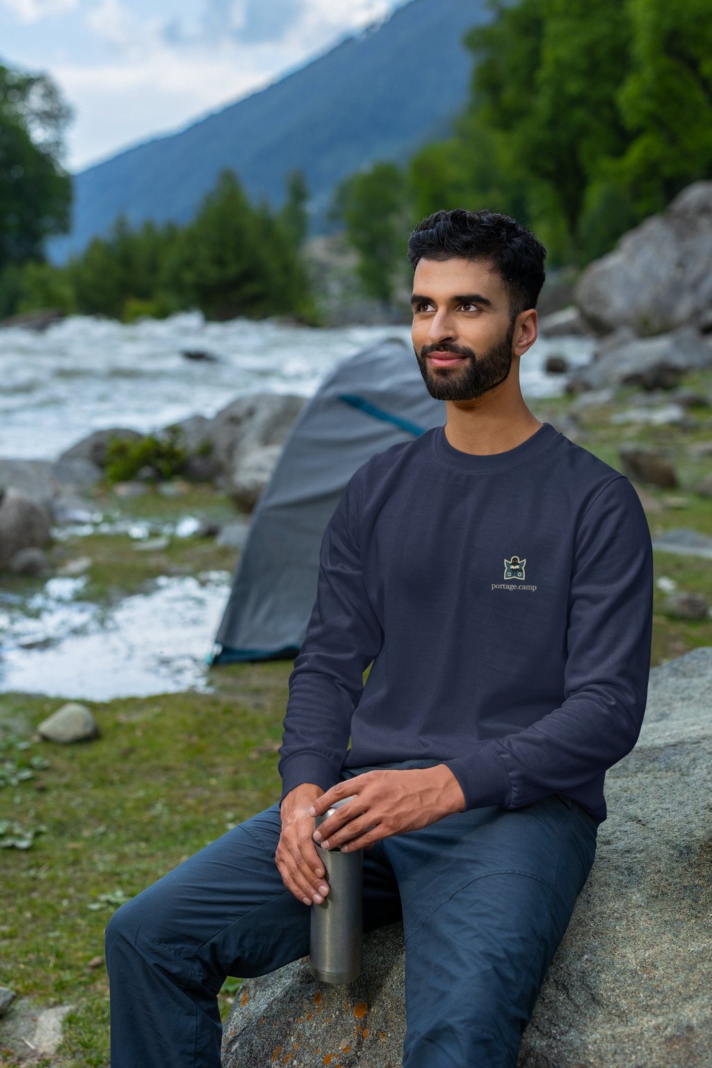 Night Library Garment-Dyed Crewneck in True Navy, worn by a man seated on a rock outdoors, with the Night Library logo and portage.camp on the chest. Digital product mockup.