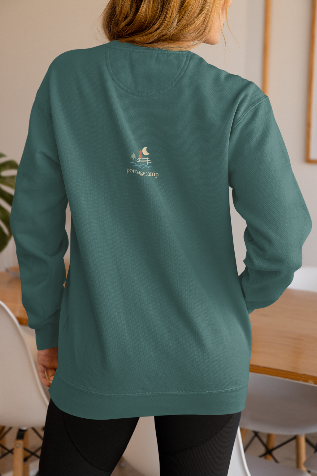 Quiet Hours Garment-Dyed Crewneck (Front Print) in Blue Spruce, worn by a woman seen from behind at home, showing the portage.camp emblem at the upper back. Digital product mockup.