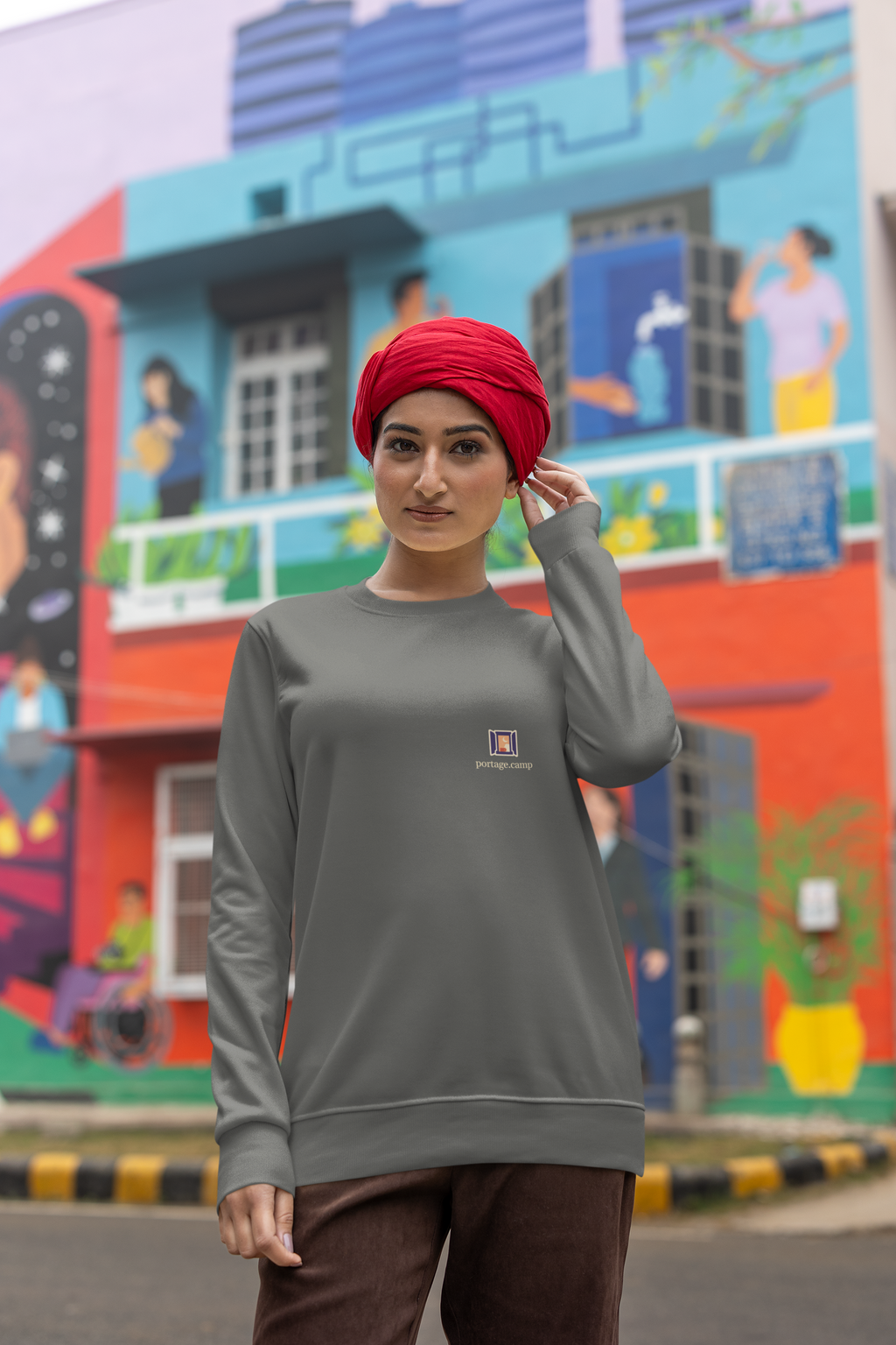 Open Late Garment-Dyed Crewneck in Pepper, worn by a woman in a turban on a busy street, with the Open Late logo and portage.camp on the chest. Digital product mockup.