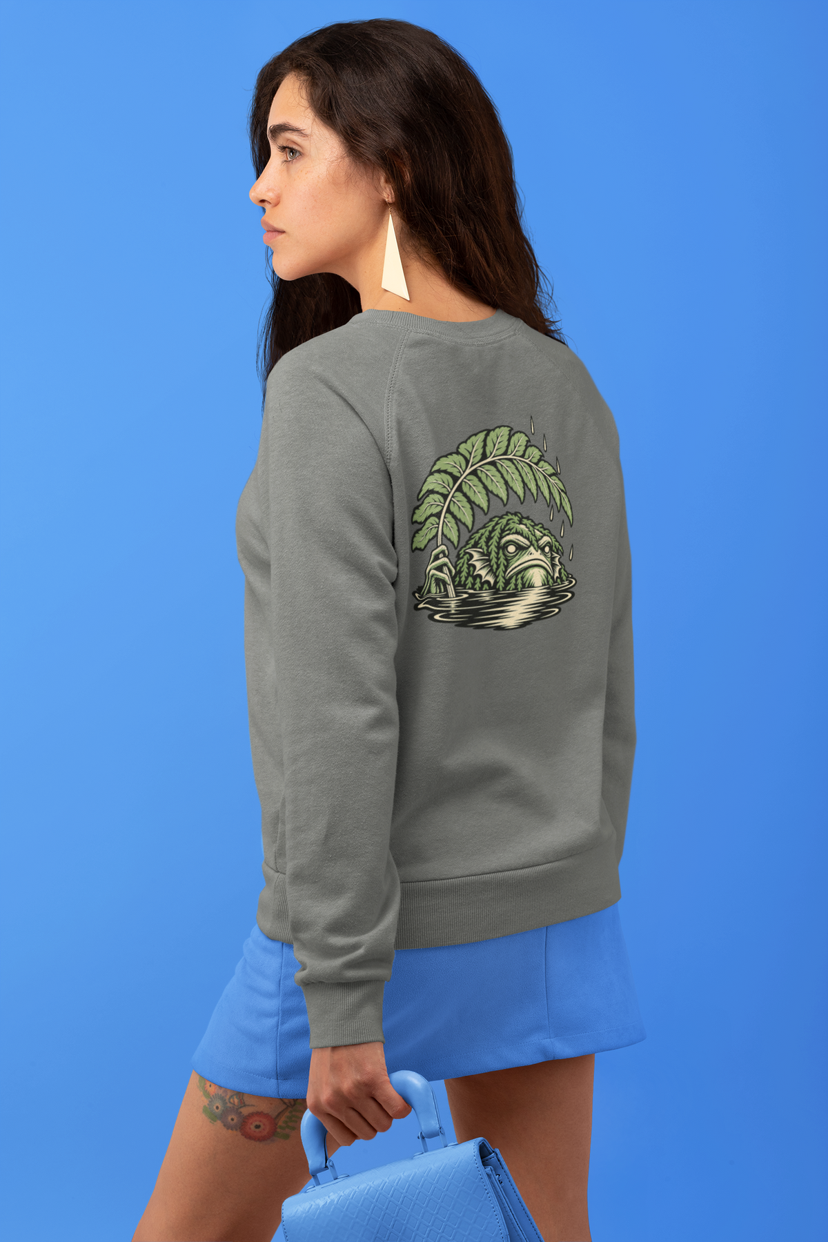 Rain Check Garment-Dyed Crewneck in Pepper, worn by a woman seen from behind against a blue backdrop, showing the large print on the back. Digital product mockup.