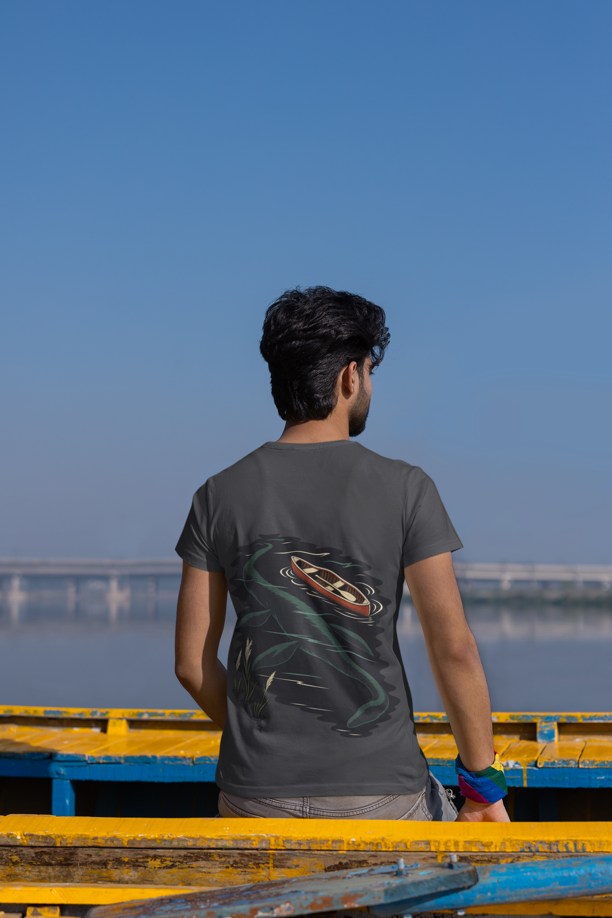 Seen Briefly Garment-Dyed Tee in Navy, worn by a man seen from behind seated on a canoe by the water, showing the large print on the back. Digital product mockup.