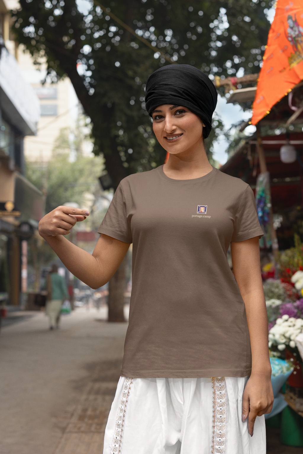 Open Late Garment-Dyed Tee in Espresso, worn by a woman in a turban by a flower market, with the Open Late logo and portage.camp on the chest. Digital product mockup.
