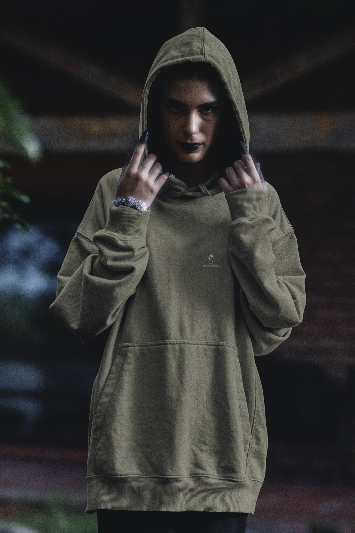 Small Hours Heavyweight Hoodie in Olive, worn by a woman in a dark forest, with the Small Hours logo and portage.camp on the left chest. Digital product mockup.