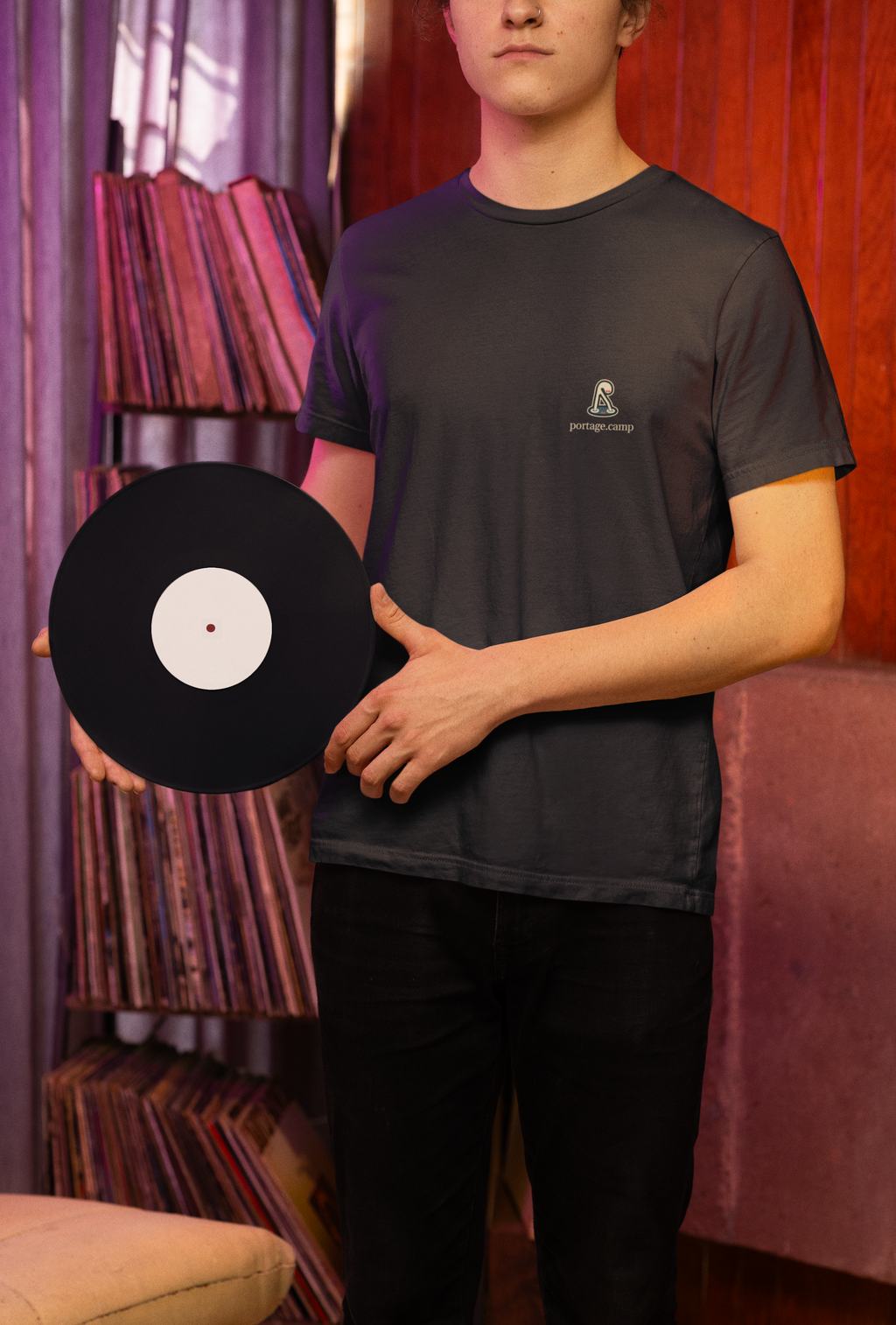Small Hours Garment-Dyed Tee in Black, worn by a man holding a vinyl record in a record shop, with the Small Hours logo and portage.camp on the chest. Digital product mockup.