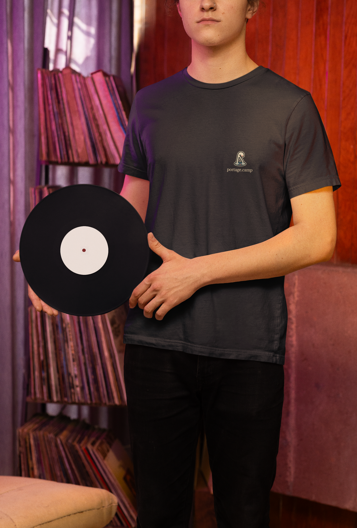 Small Hours Garment-Dyed Tee in Black, worn by a man holding a vinyl record in a record shop, with the Small Hours logo and portage.camp on the chest. Digital product mockup.