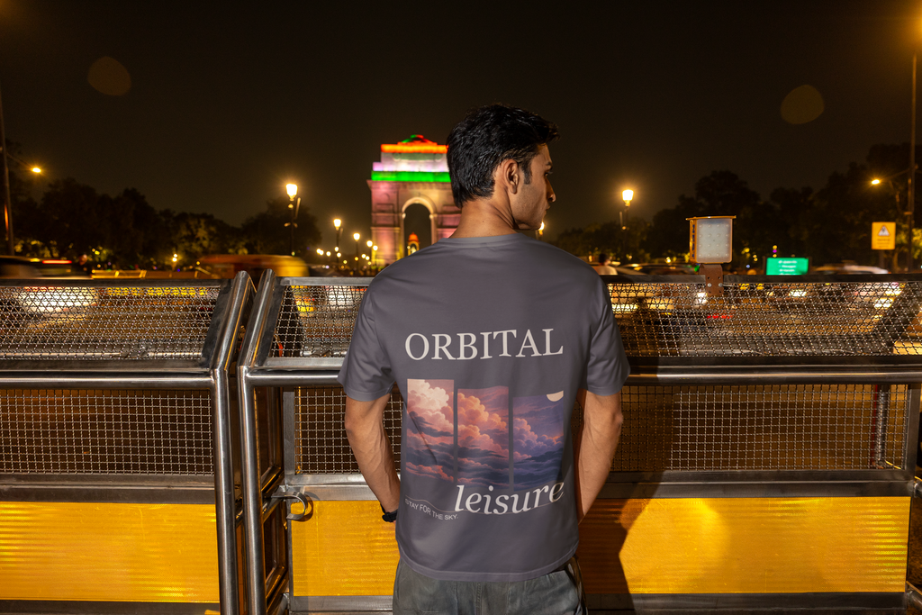 Stay for the Sky Garment-Dyed Tee in Graphite, worn by a man seen from behind on a bridge over the city at night, showing the large print on the back. Digital product mockup.