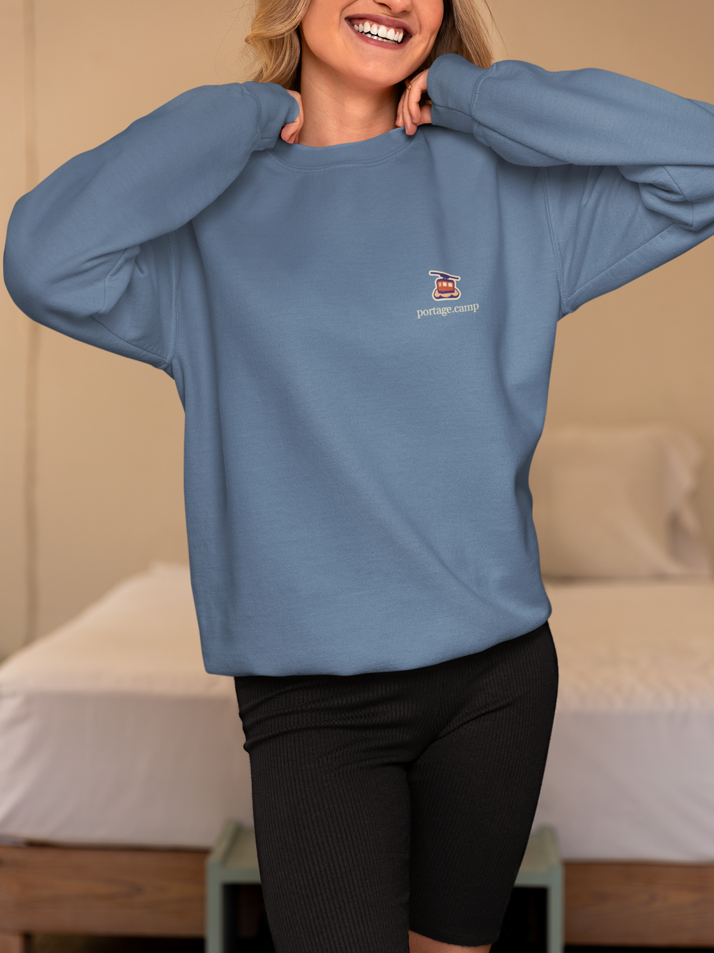 Cloud Ferry Garment-Dyed Crewneck in Blue Jean, worn by a woman in a bedroom, with the Cloud Ferry logo and portage.camp on the chest. Digital product mockup.