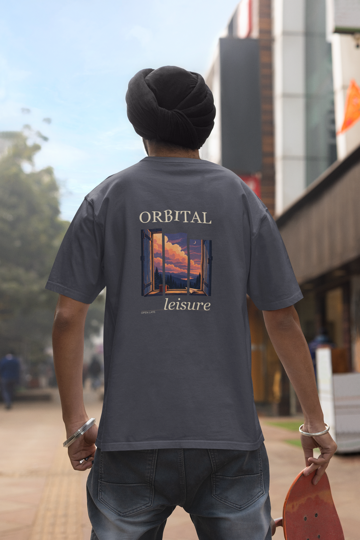 Open Late Garment-Dyed Tee in Navy, worn by a man seen from behind in a turban on a busy street, showing the large print on the back. Digital product mockup.
