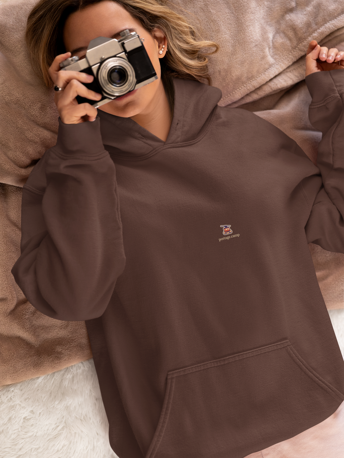 Cloud Ferry Heavyweight Hoodie in Brown, worn by a woman in bed with a film camera, with the Cloud Ferry logo and portage.camp on the left chest. Digital product mockup.