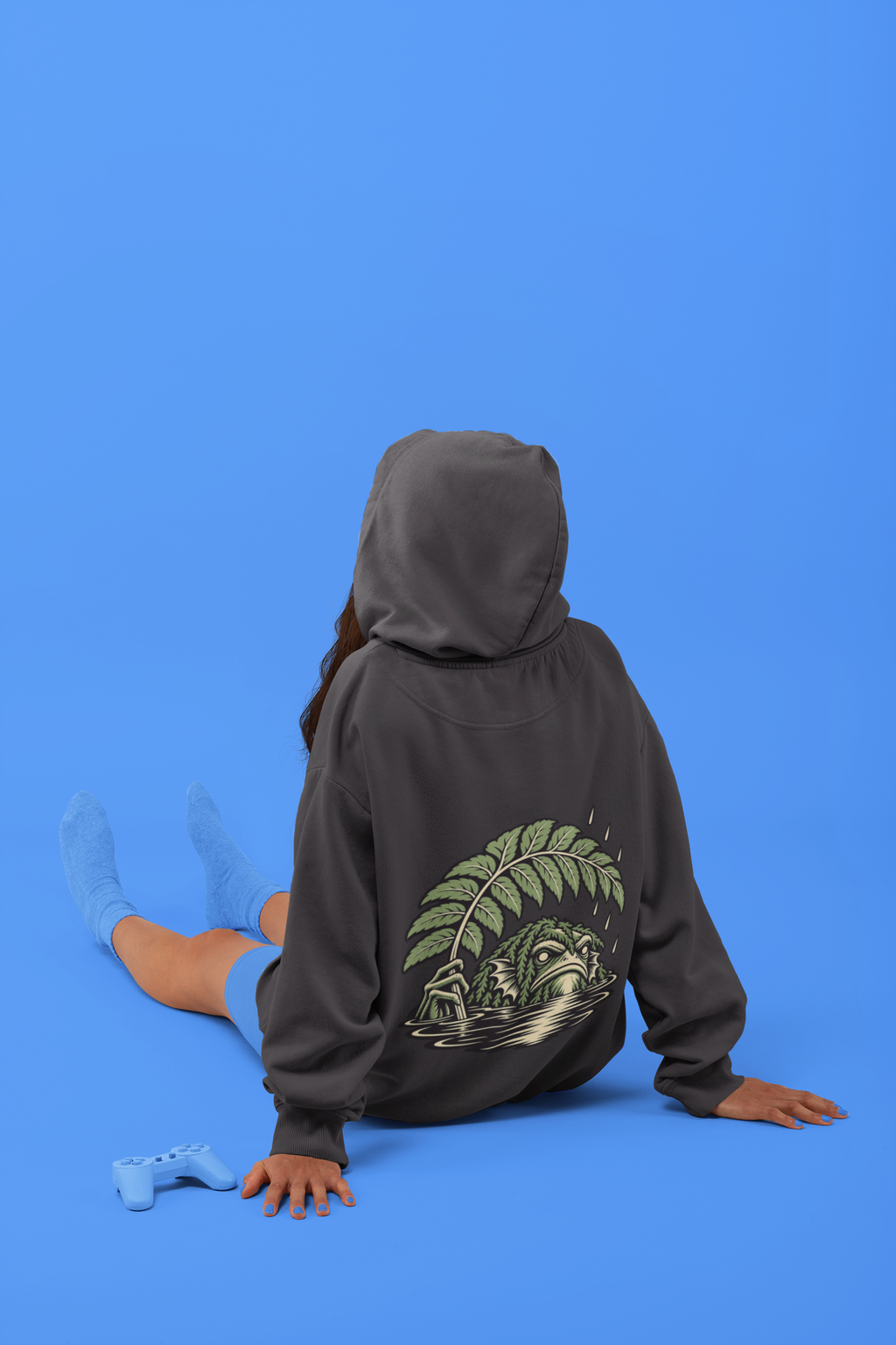 Rain Check Midweight Hoodie in Black, worn by a woman seen from behind seated on a studio floor, showing the large print on the back. Digital product mockup.