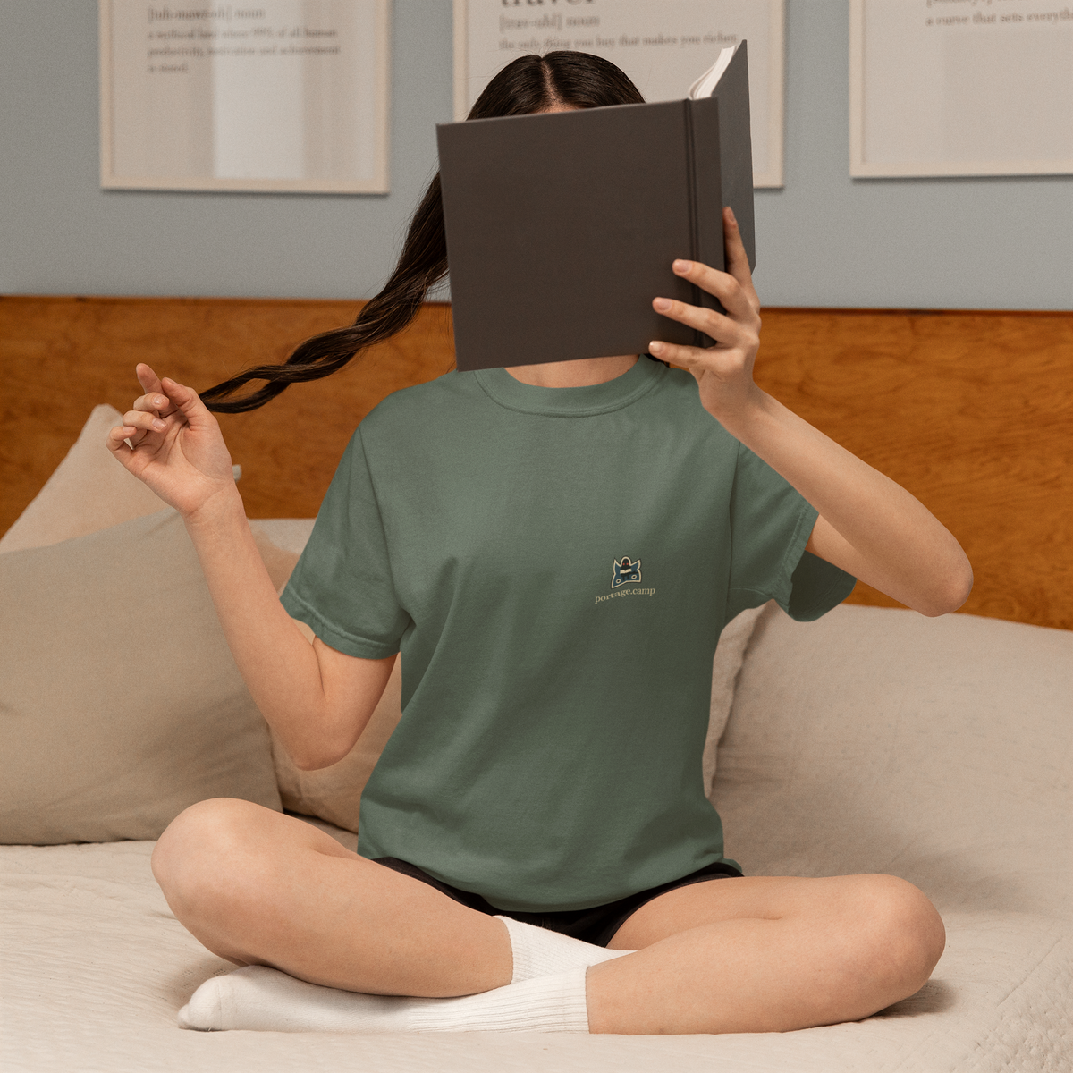 Night Library Garment-Dyed Tee in Blue Spruce, worn by a woman at home, hiding behind a book, with the Night Library logo and portage.camp on the chest. Digital product mockup.