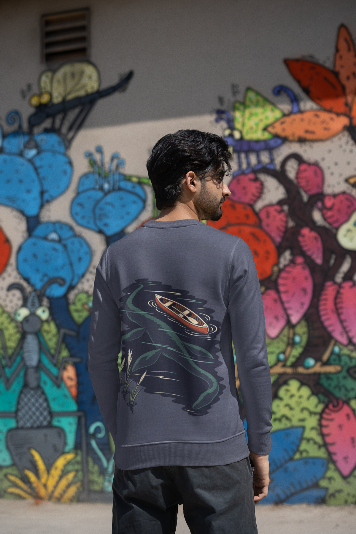 Seen Briefly Garment-Dyed Crewneck in Denim, worn by a man seen from behind by a painted wall in the city, showing the large print on the back. Digital product mockup.