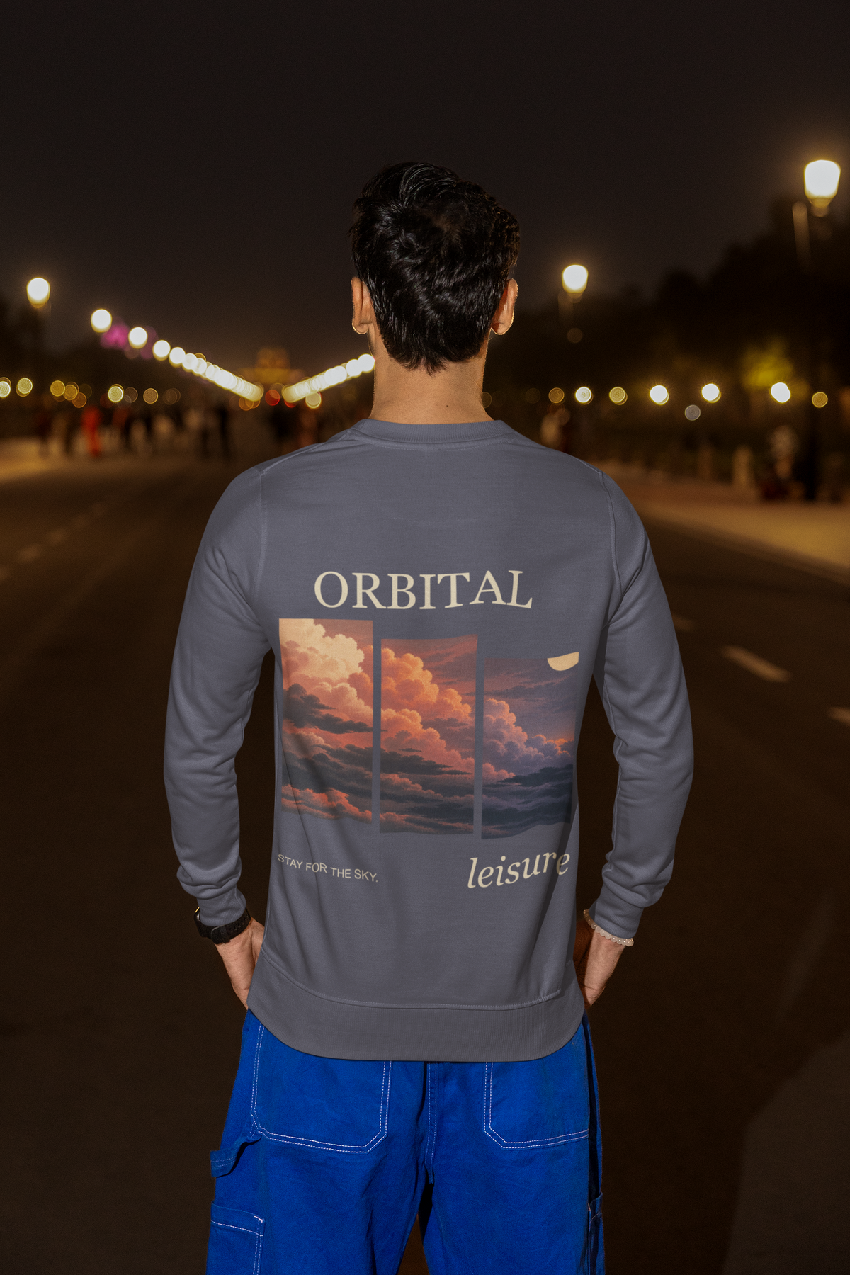 Stay for the Sky Garment-Dyed Crewneck in Denim, worn by a man seen from behind on a city street at night, showing the large print on the back. Digital product mockup.