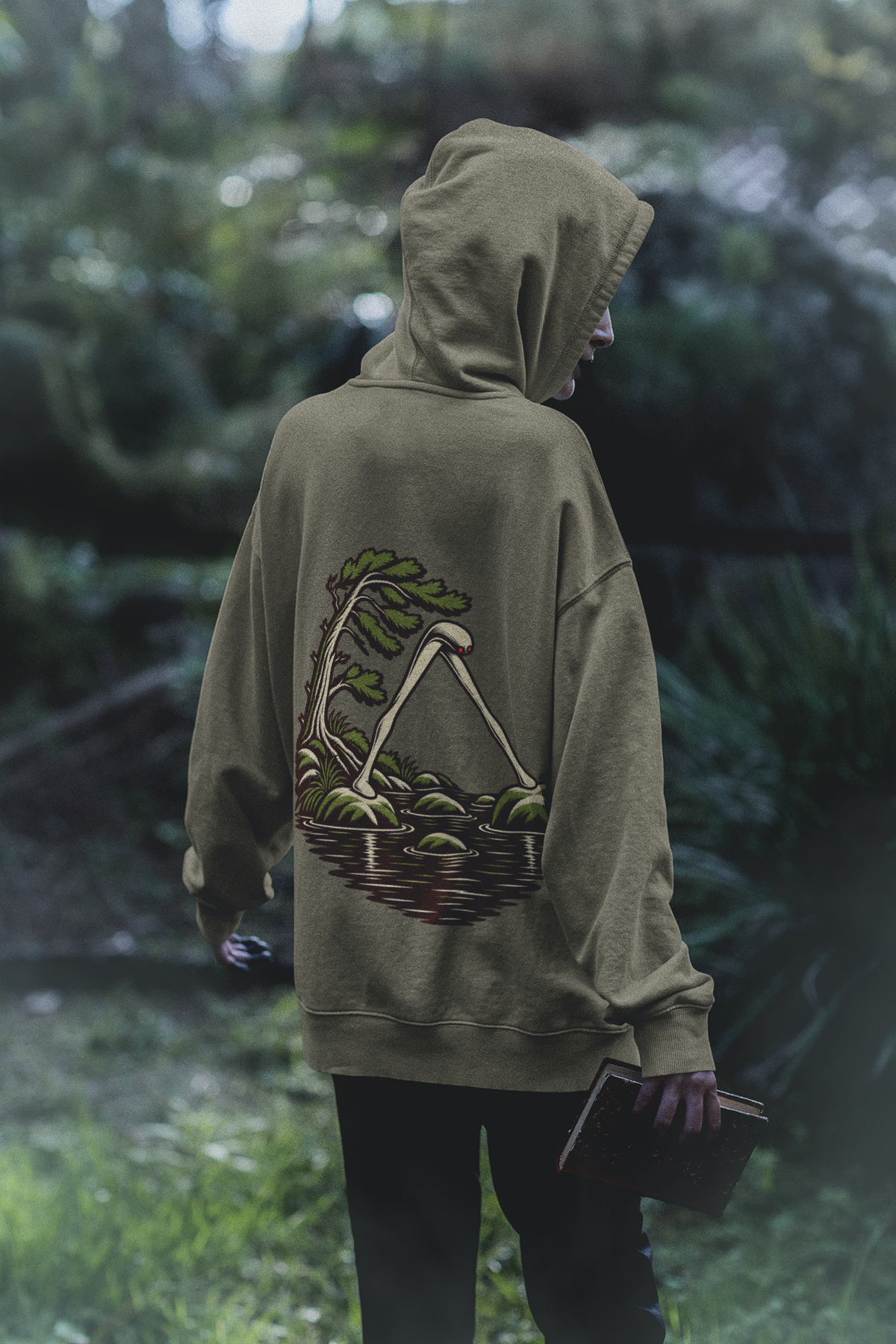 Small Hours Heavyweight Hoodie in Olive, worn by a woman seen from behind in a dark forest, showing the large print on the back. Digital product mockup.