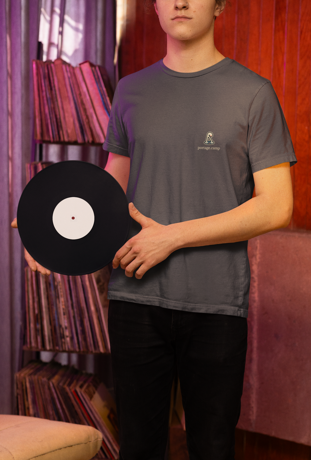 Small Hours Garment-Dyed Tee in Graphite, worn by a man holding a vinyl record in a record shop, with the Small Hours logo and portage.camp on the chest. Digital product mockup.