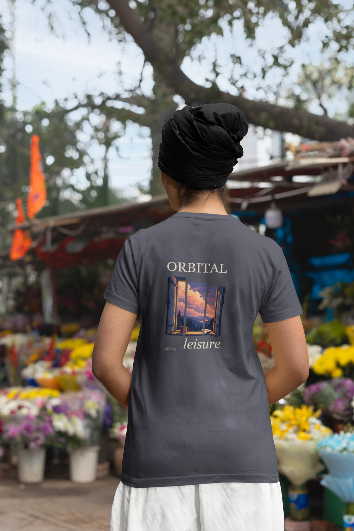 Open Late Garment-Dyed Tee in Navy, worn by a woman seen from behind in a turban by a flower market, showing the large print on the back. Digital product mockup.