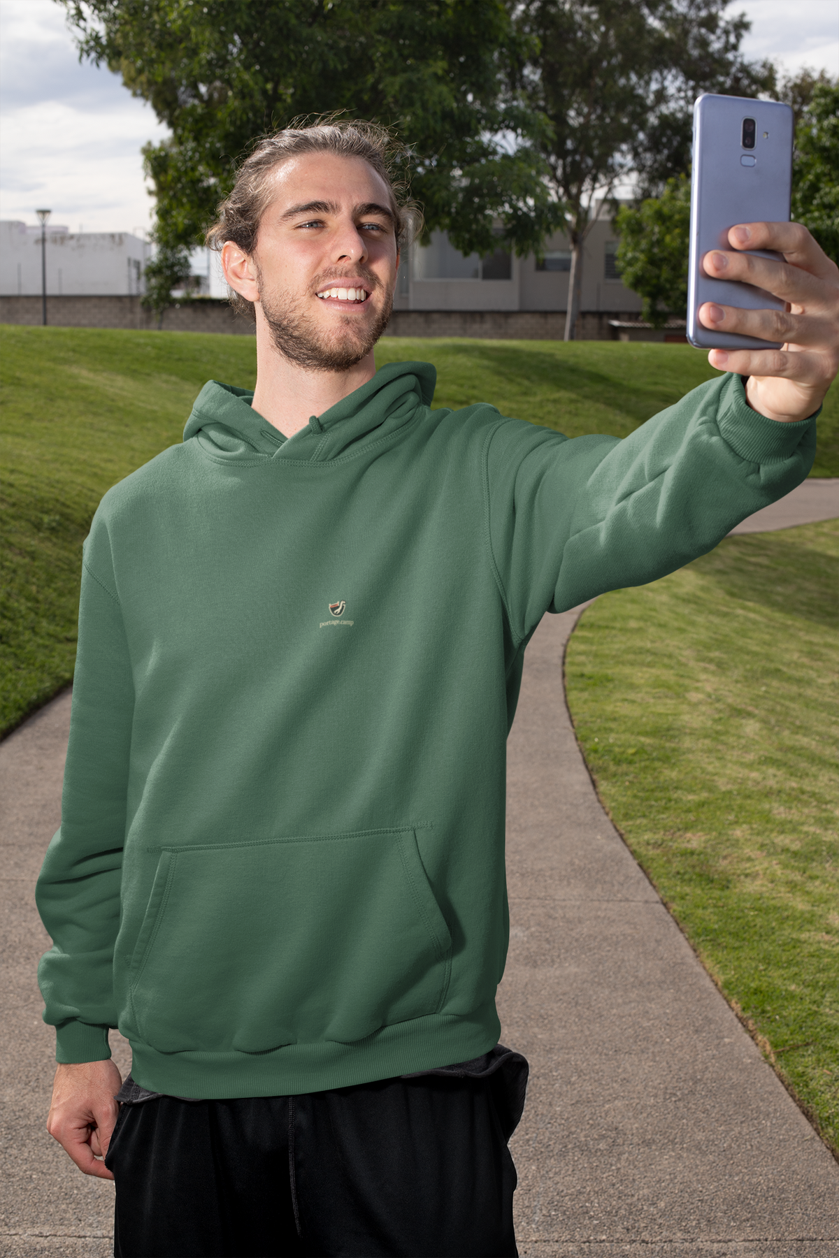 Seen Briefly Midweight Hoodie in Alpine Green, worn by a man taking a selfie on a city street, with the Seen Briefly logo and portage.camp on the chest. Digital product mockup.