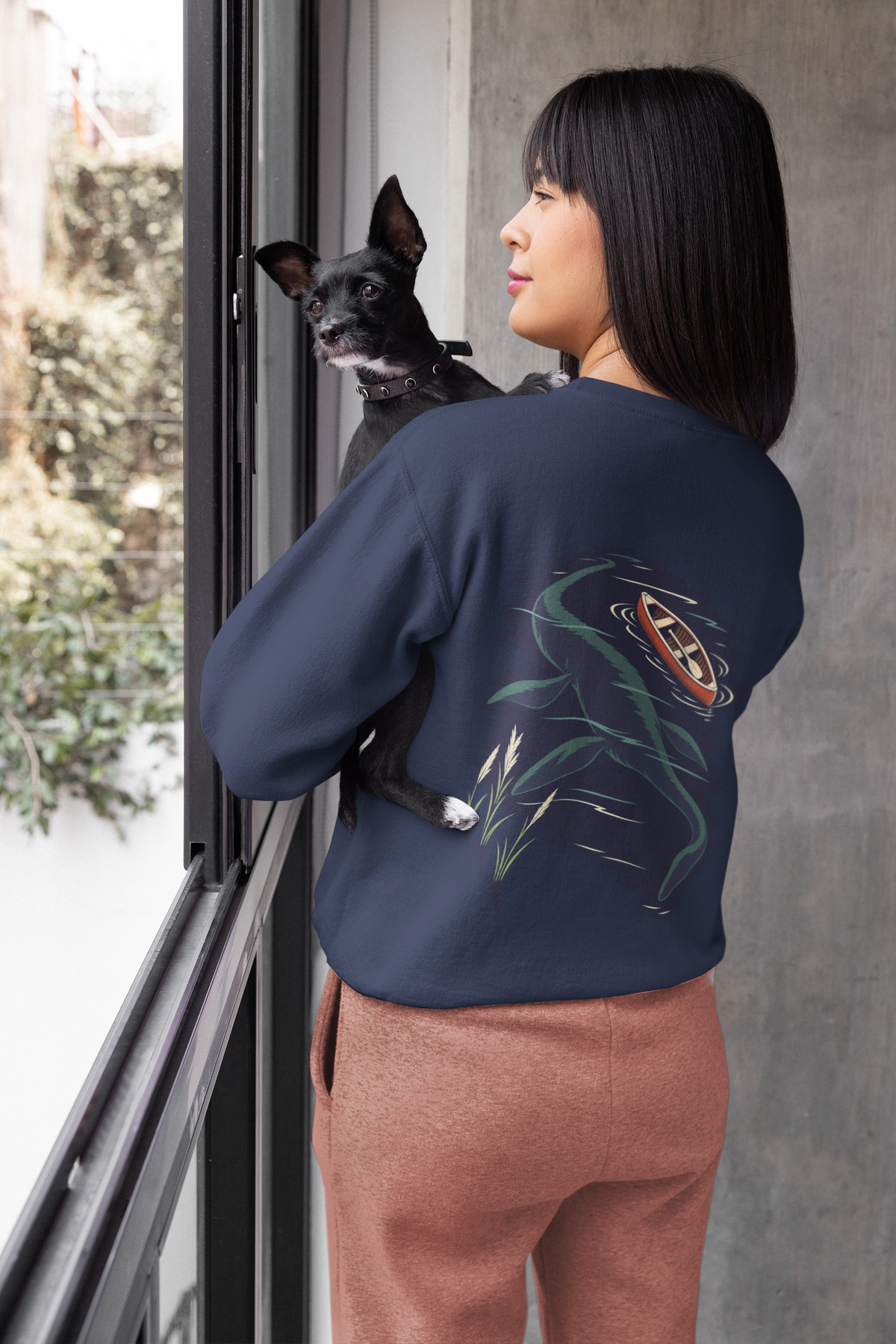 Seen Briefly Garment-Dyed Crewneck in True Navy, worn by a woman seen from behind at home, holding her dog, showing the large print on the back. Digital product mockup.