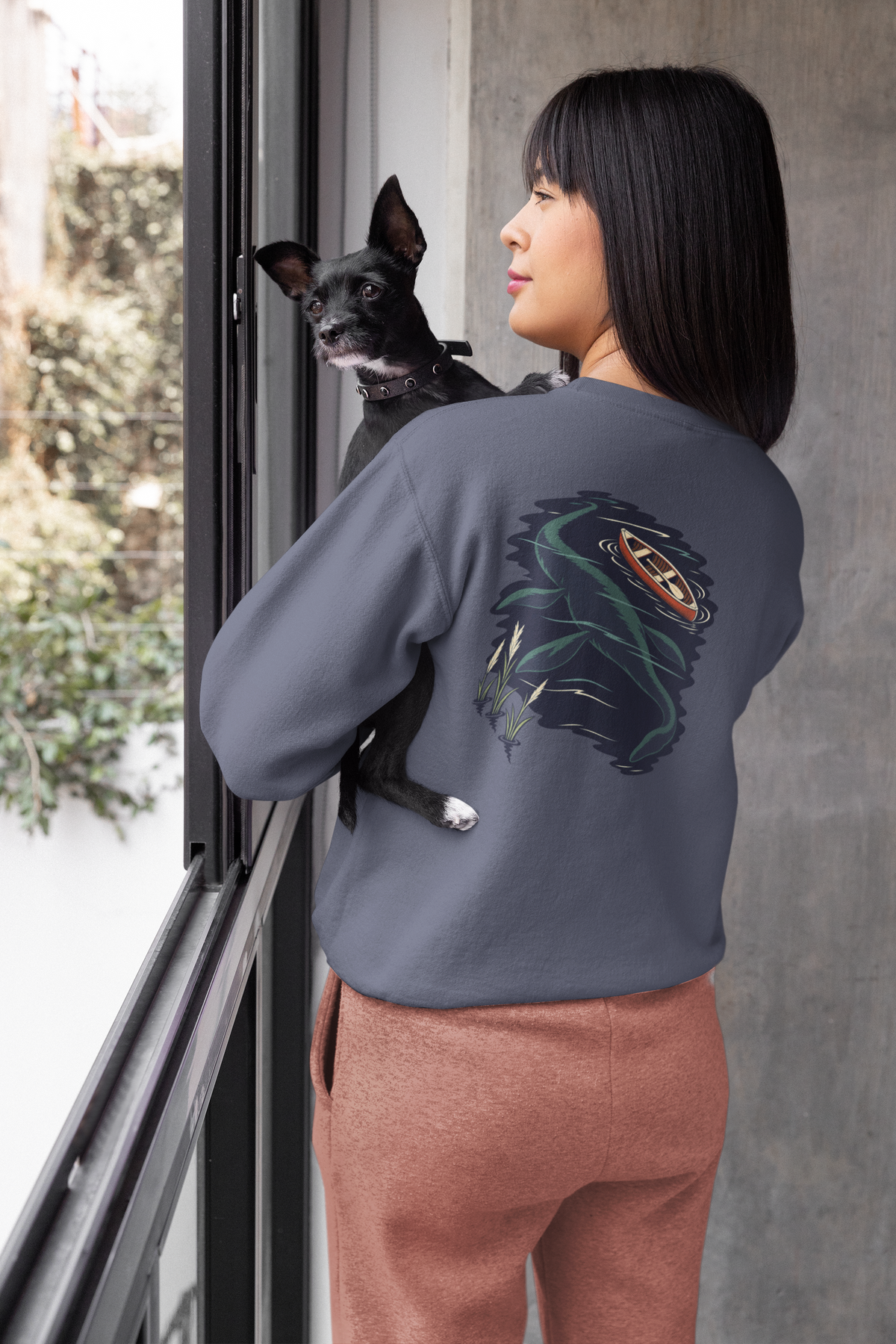 Seen Briefly Garment-Dyed Crewneck in Denim, worn by a woman seen from behind at home, holding her dog, showing the large print on the back. Digital product mockup.