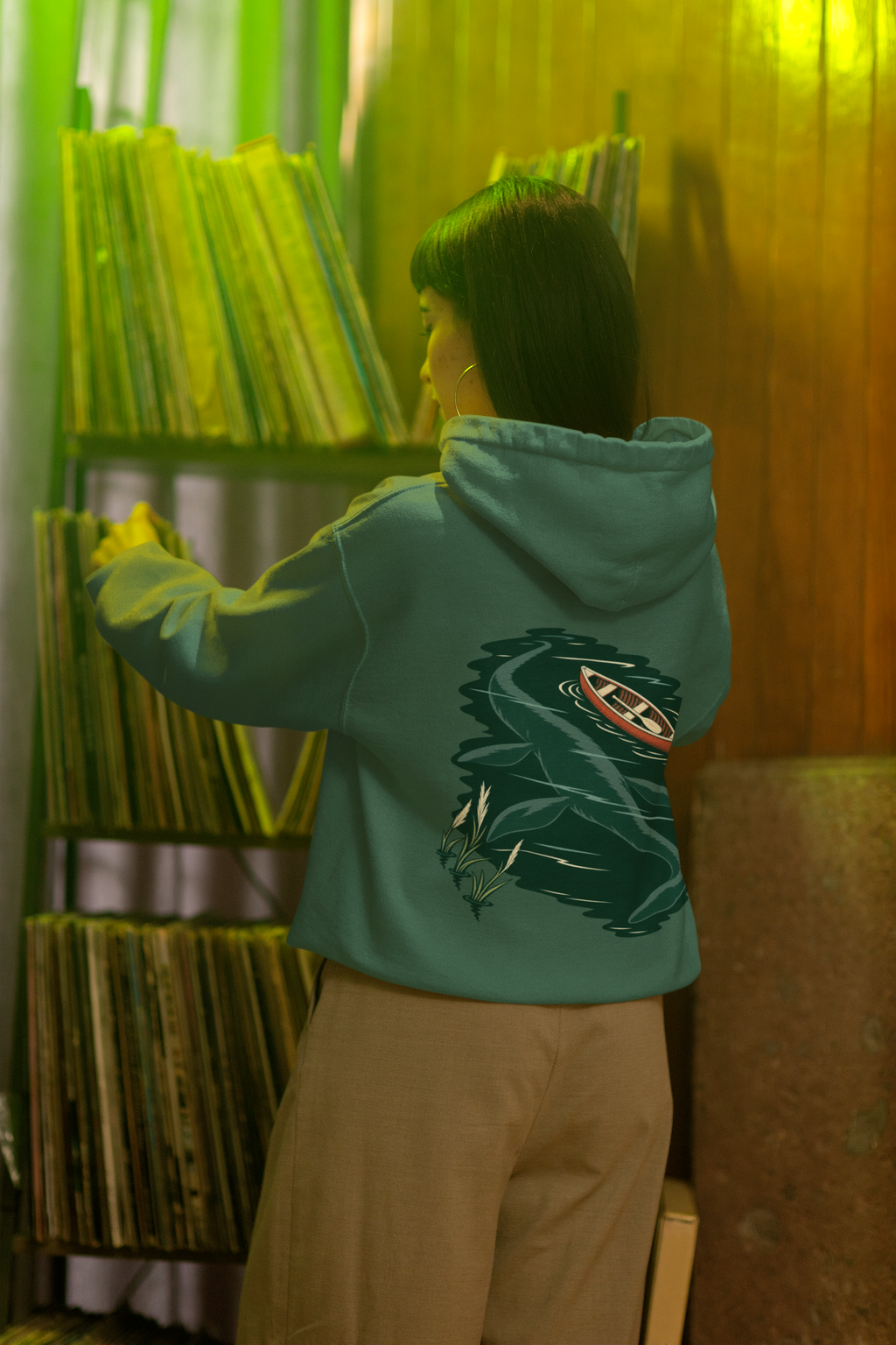 Seen Briefly Midweight Hoodie in Alpine Green, worn by a woman seen from behind in a vintage record shop, showing the large print on the back. Digital product mockup.