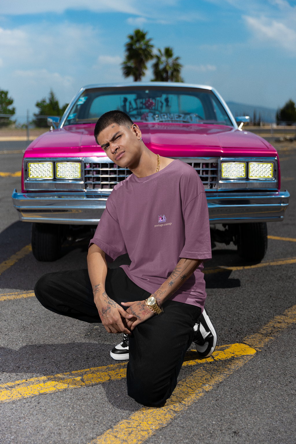 Amethyst After Hours Garment-Dyed Tee in Berry, worn by a man crouched in front of a pink classic car, with the Amethyst After Hours logo and portage.camp on the chest. Digital product mockup.