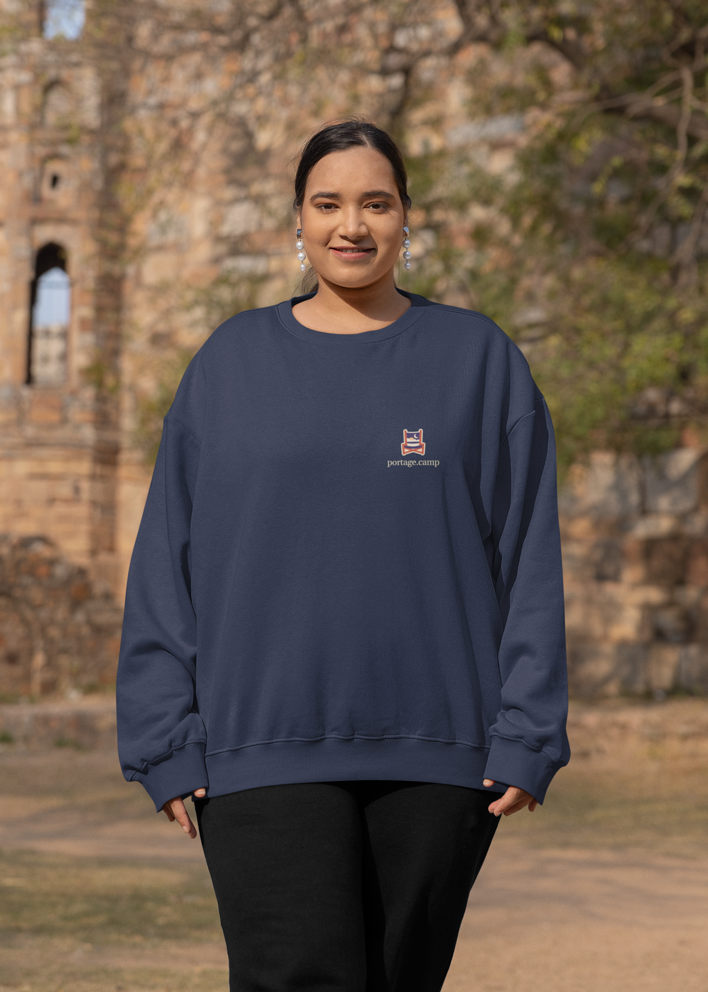 Edge of Evening Garment-Dyed Crewneck in True Navy, worn by a woman by an old stone building, with the Edge of Evening logo and portage.camp on the chest. Digital product mockup.
