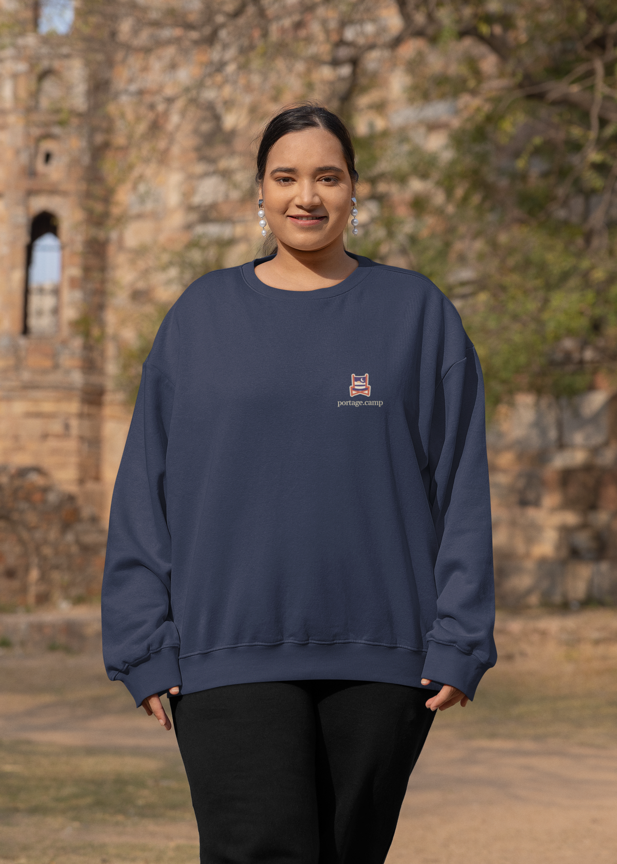 Edge of Evening Garment-Dyed Crewneck in True Navy, worn by a woman by an old stone building, with the Edge of Evening logo and portage.camp on the chest. Digital product mockup.