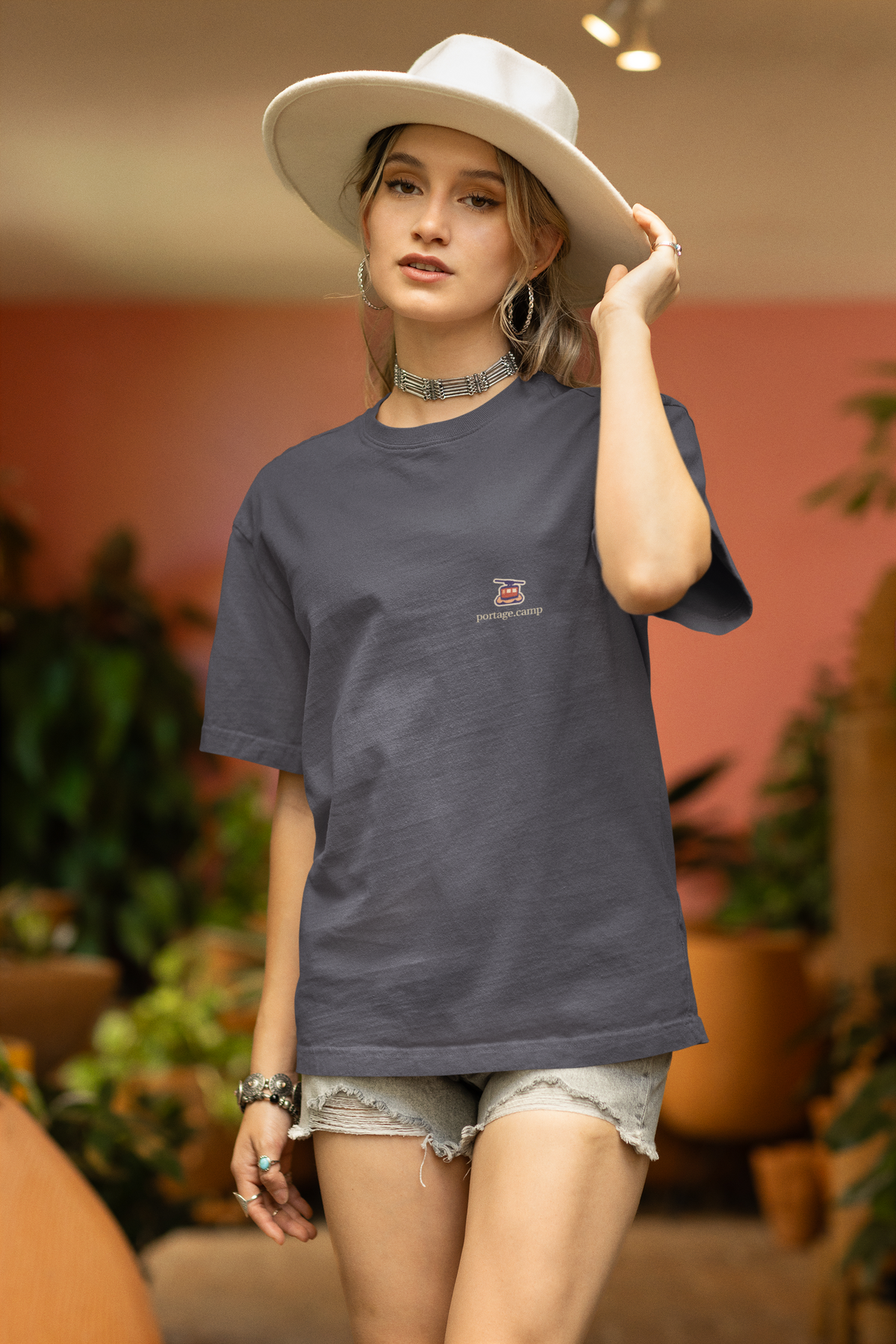 Cloud Ferry Garment-Dyed Tee in Navy, worn by a woman in a sun hat among potted plants, with the Cloud Ferry logo and portage.camp on the chest. Digital product mockup.