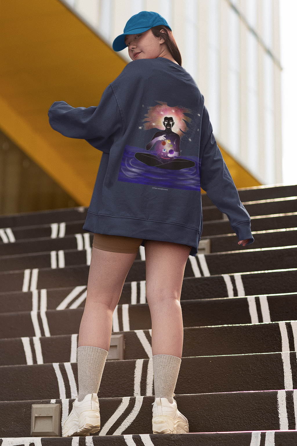 Orbit Keeper Garment-Dyed Crewneck in True Navy, worn by a woman seen from behind on outdoor stairs, showing the large print on the back. Digital product mockup.