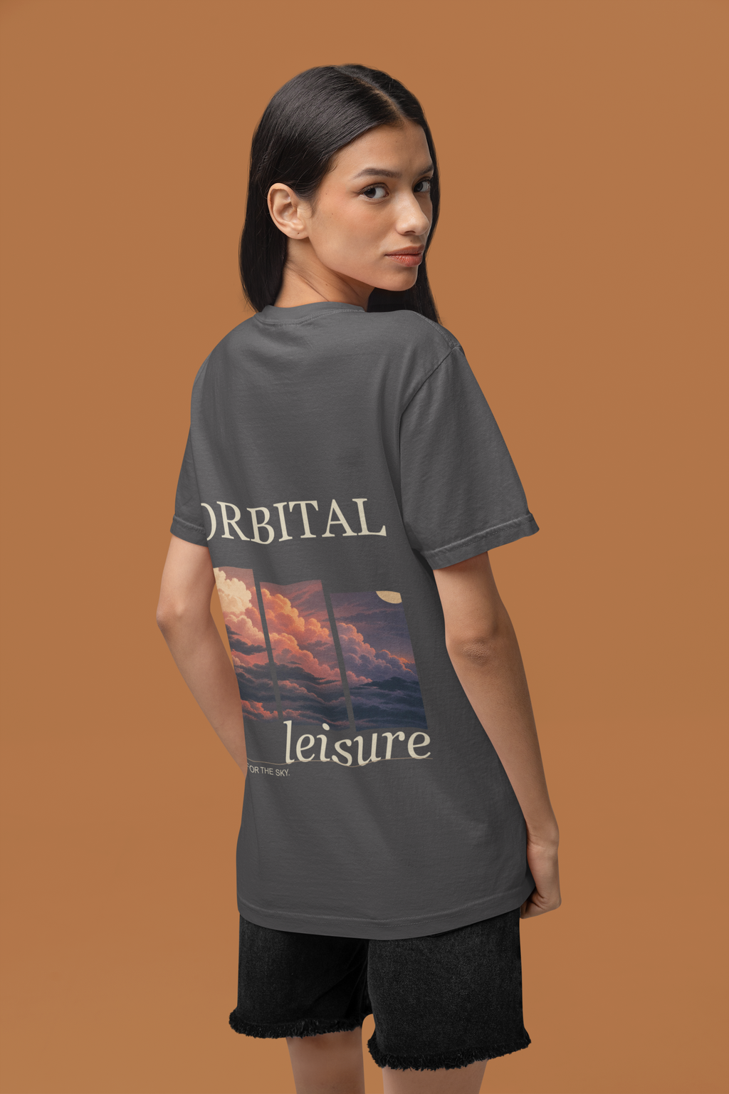 Stay for the Sky Garment-Dyed Tee in Graphite, worn by a woman seen from behind in a studio against a warm brown wall, showing the large print on the back. Digital product mockup.