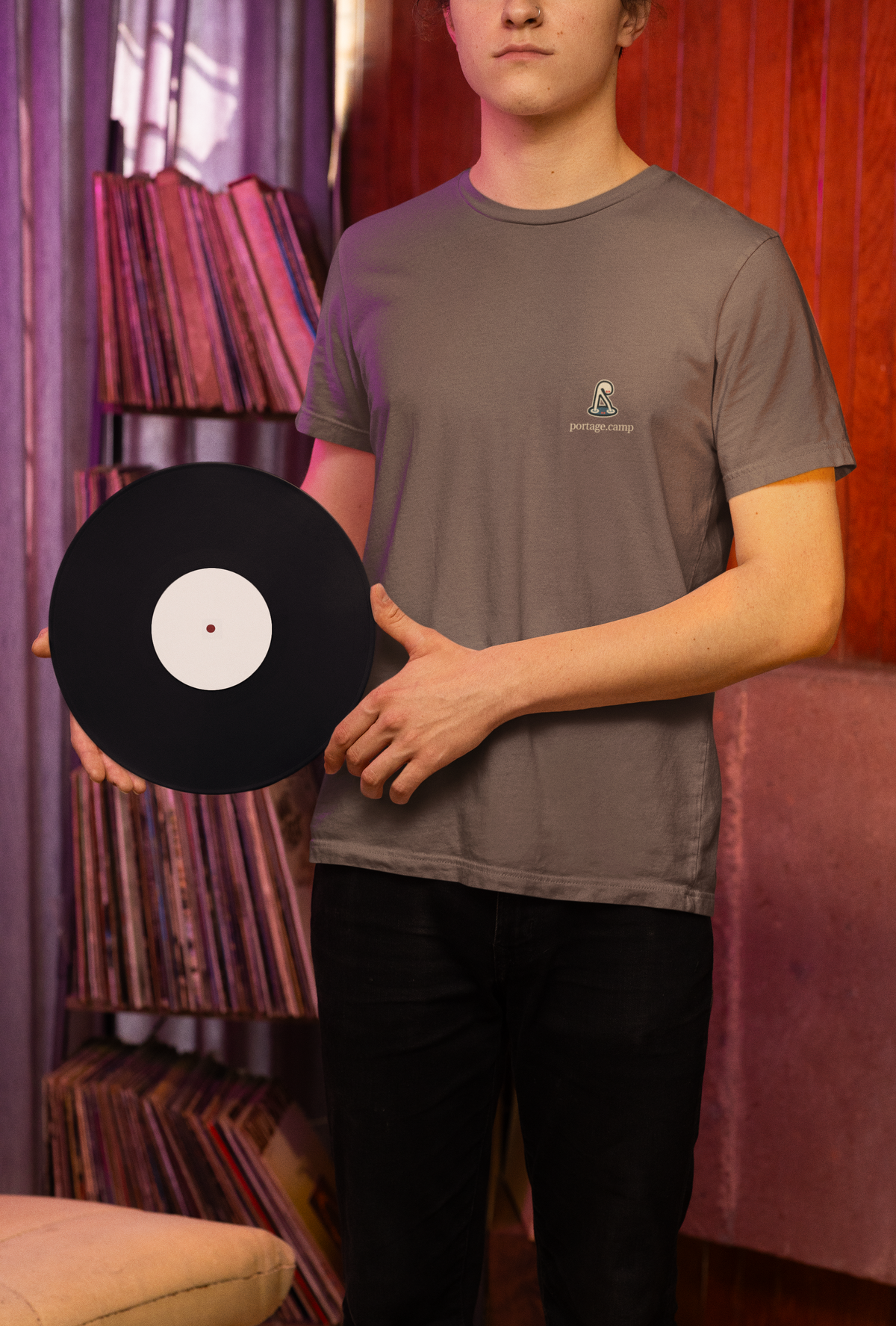 Small Hours Garment-Dyed Tee in Espresso, worn by a man holding a vinyl record in a record shop, with the Small Hours logo and portage.camp on the chest. Digital product mockup.