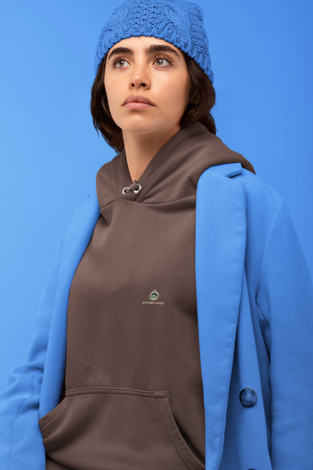Rain Check Heavyweight Hoodie in Brown, worn by a woman against a blue backdrop, with the Rain Check logo and portage.camp on the left chest. Digital product mockup.