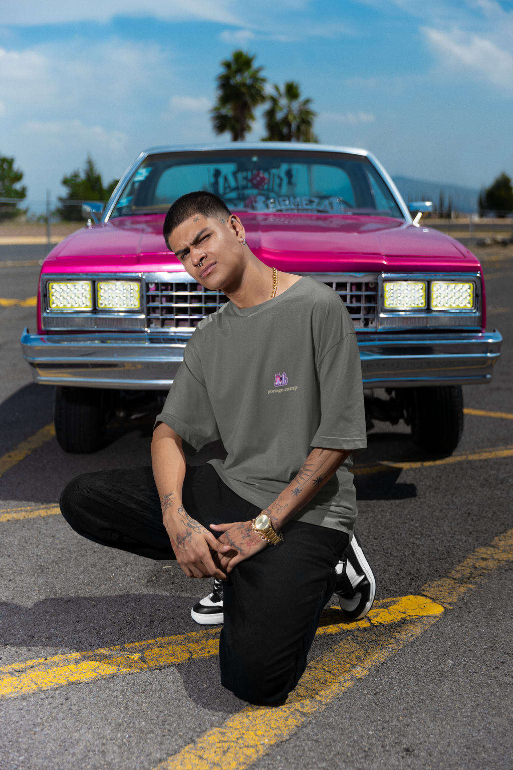 Amethyst After Hours Garment-Dyed Tee in Pepper, worn by a man crouched in front of a pink classic car, with the Amethyst After Hours logo and portage.camp on the chest. Digital product mockup.